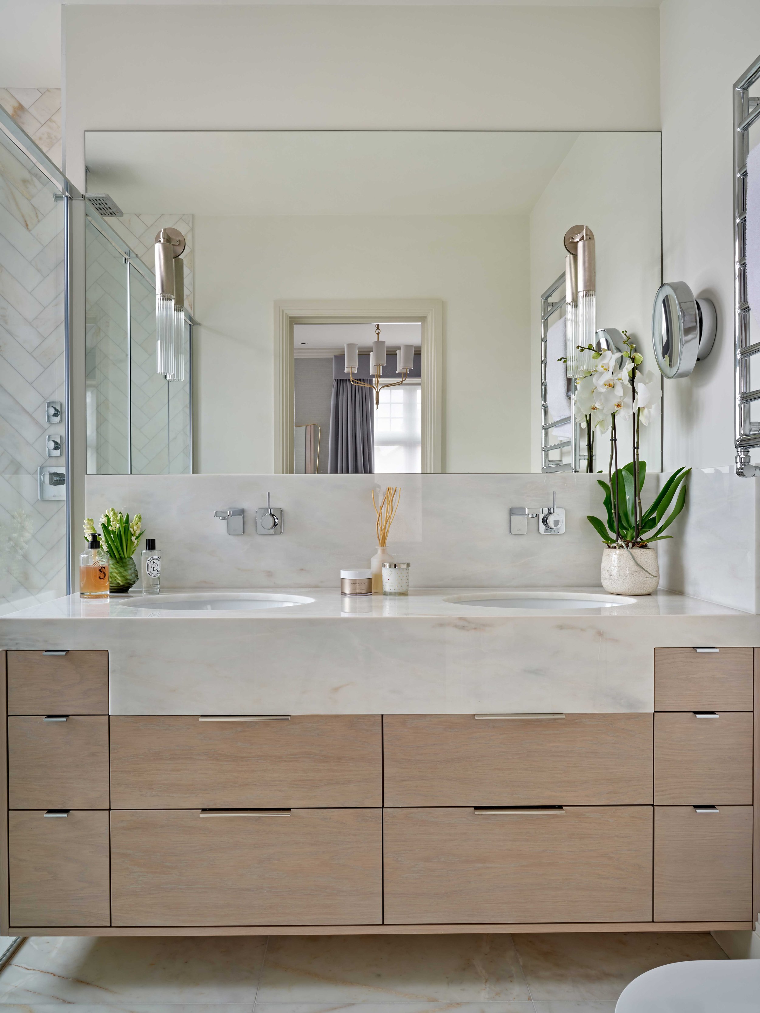 Modern bathroom vanity with a large mirror, white countertop, and wooden drawers, decorated with potted plants, candles, and toiletries, with a reflection of a doorway leading to a dining area.