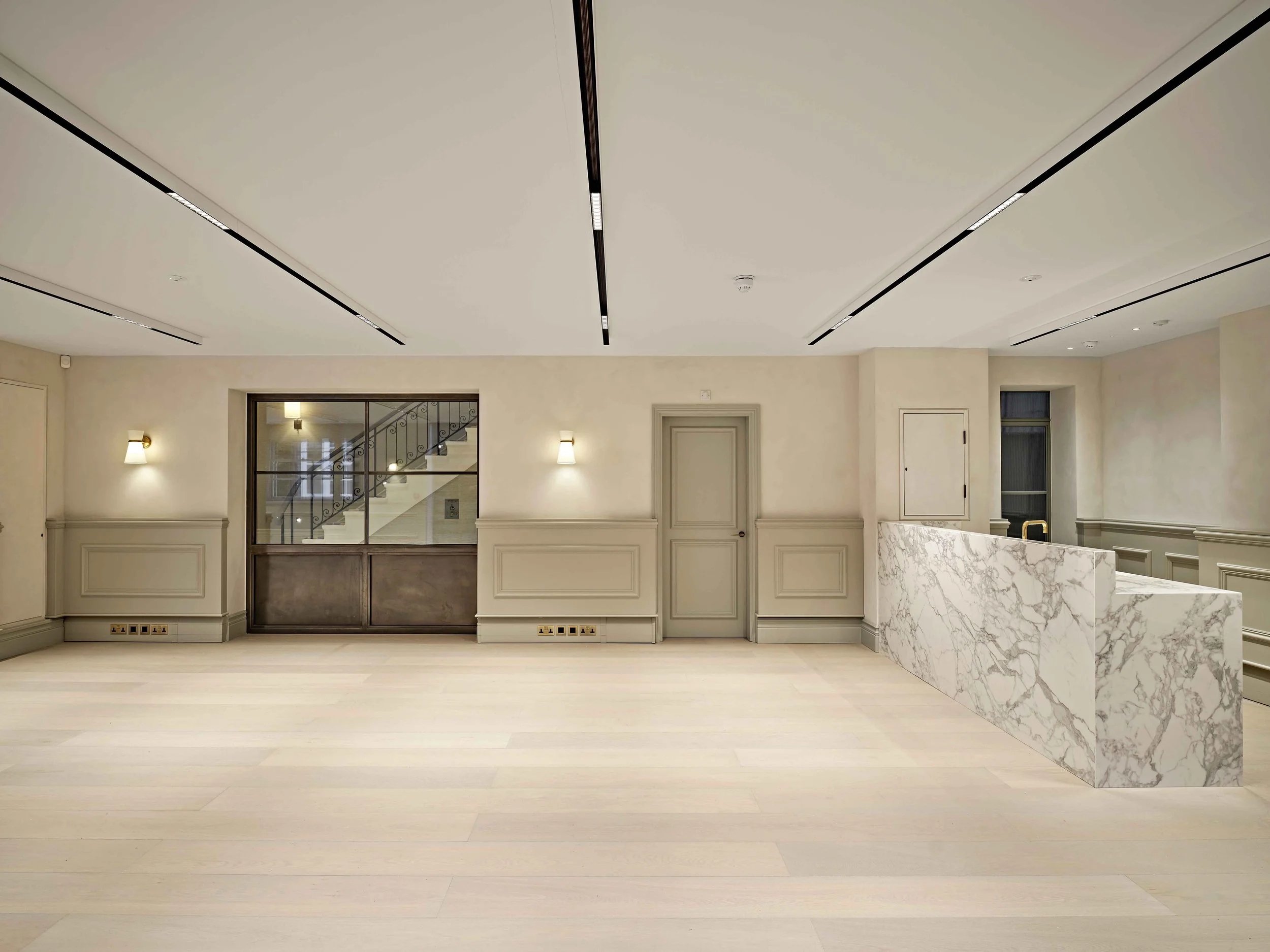 An empty, modern interior room with a high ceiling, cream-colored walls with decorative molding, light-colored wooden flooring, and a marble reception desk on the right. There are wall sconces and a staircase visible through a window in the backgroun