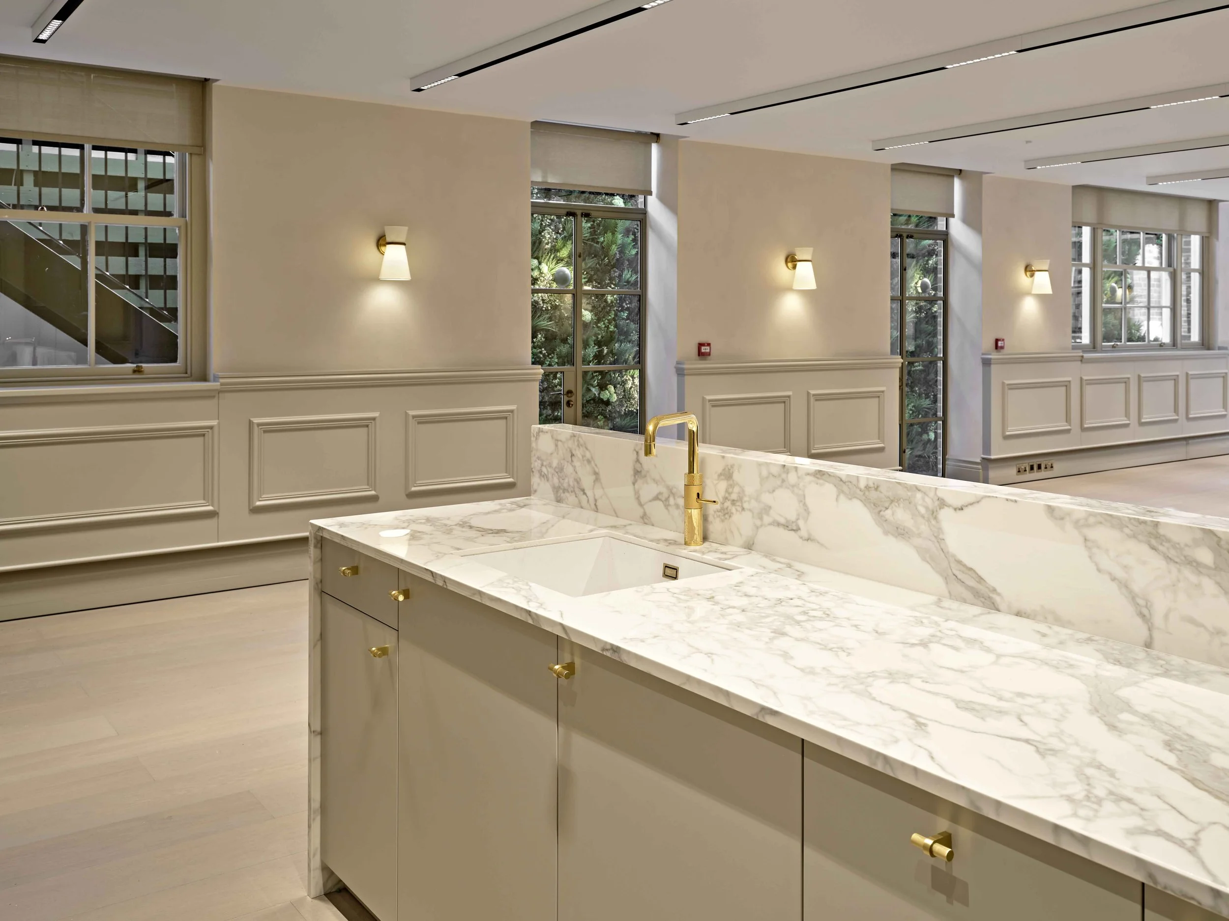 Modern kitchen with white marble island, gold fixtures, beige paneled walls, large windows with views of greenery, and ceiling lighting.