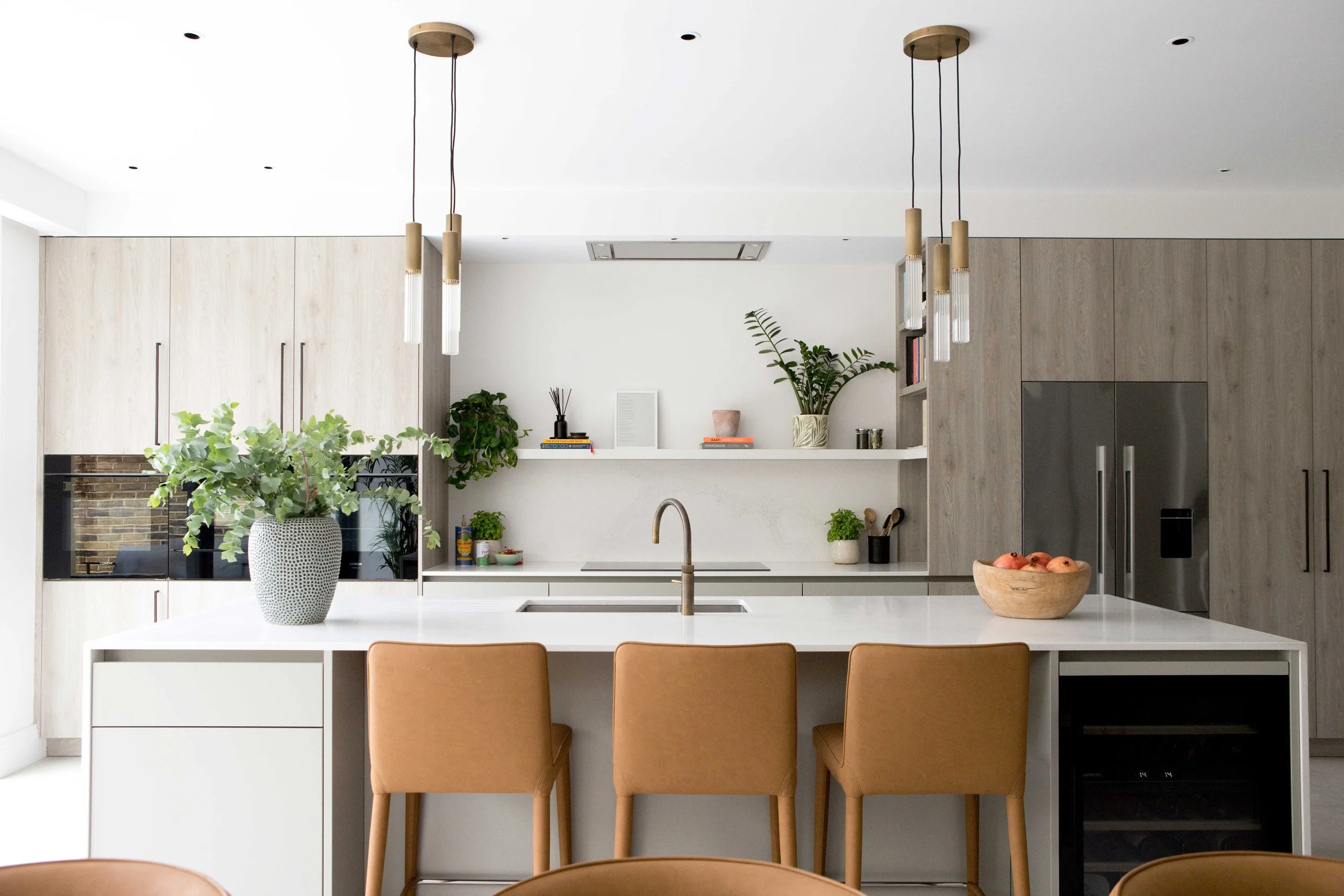 Modern kitchen with white countertop, light wood cabinets, hanging pendant lights, potted plants, and beige chairs.