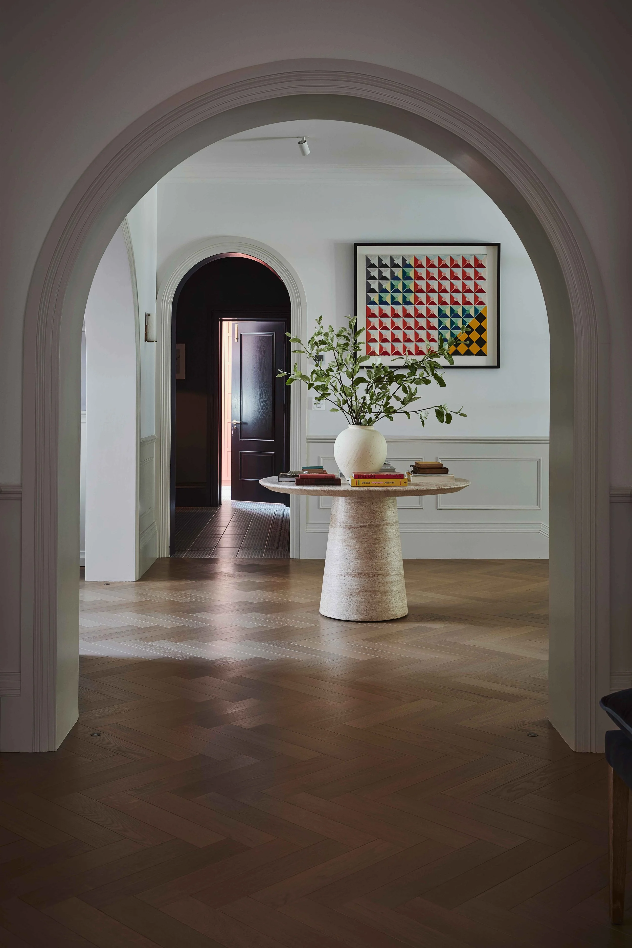 A hallway view through a rounded archway, featuring a round table with a large white vase and books, a framed geometric artwork on the wall, and an open dark door leading outside, with wood flooring and white walls.