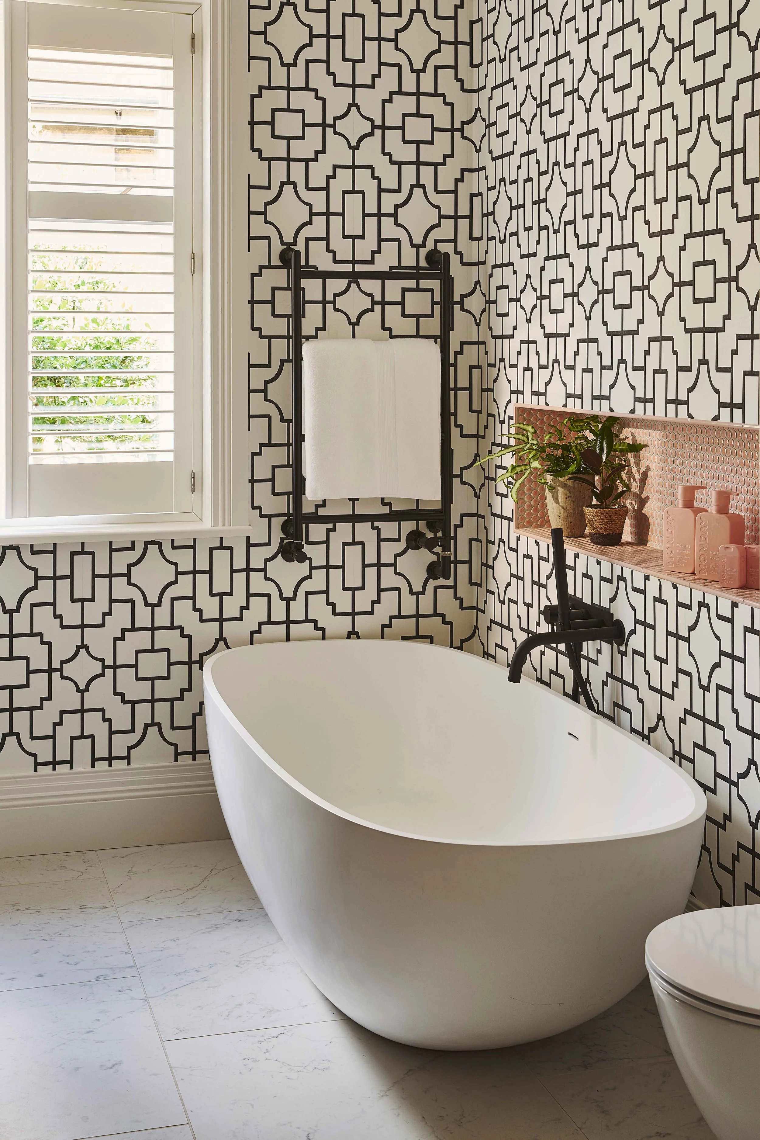 A bathroom with a modern freestanding white bathtub, black and white patterned wallpaper, a window with shutters, a towel rack with White towel, a shelf with plants and pink soap bottles, and a marble tile floor.