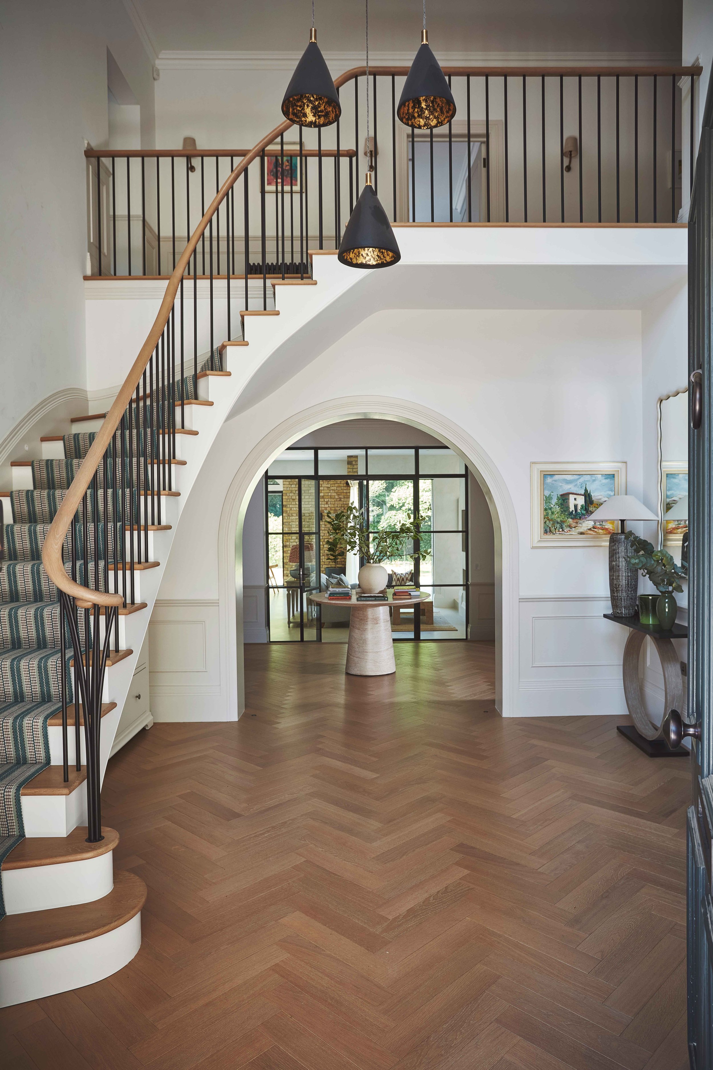 Interior view of a modern home with a staircase, black pendant lights hanging from the ceiling, and a large archway leading to another room with large windows and a table with decorative items. Artwork is hanging on the walls, and hardwood flooring is present throughout.