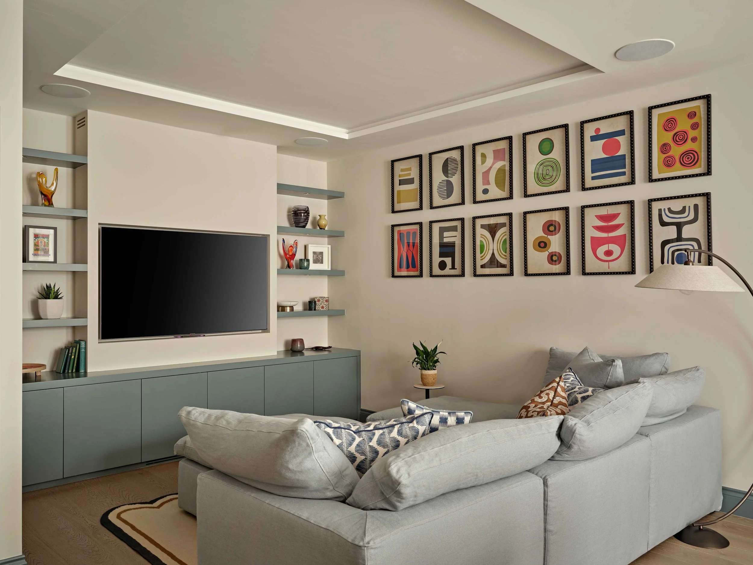 Living room with a gray couch, a small side table, a wall with framed abstract art, built-in shelves with decorative items, and a large flat-screen TV.