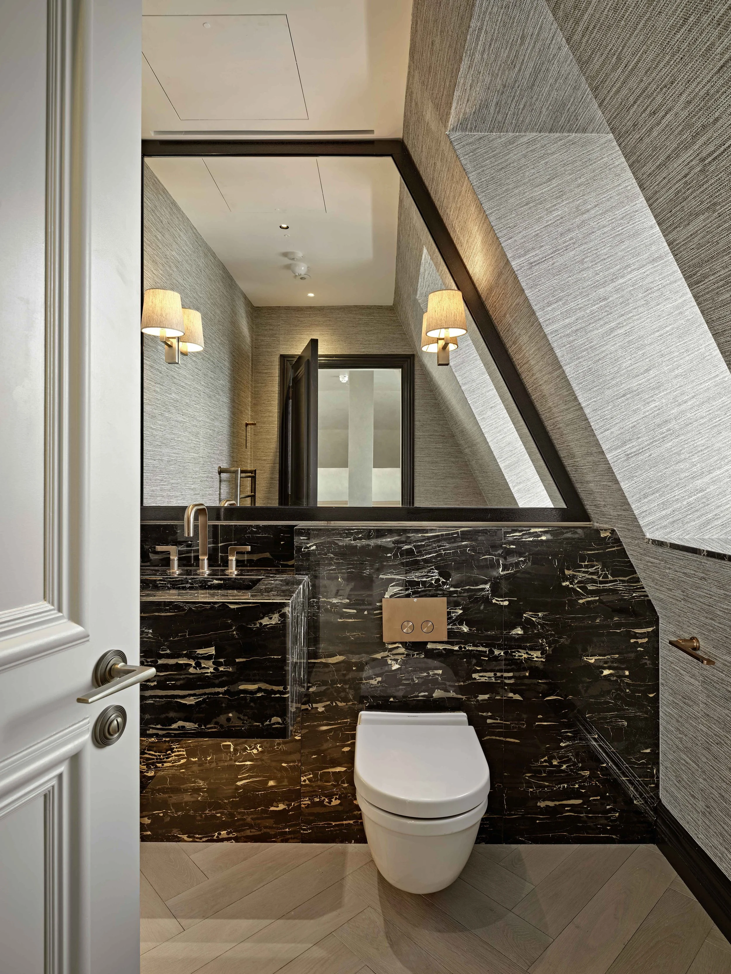 Modern bathroom featuring a wall-mounted toilet, black marble countertop with a sink, beige tile flooring, textured beige wallpaper, wall-mounted lamps, and a large mirror.