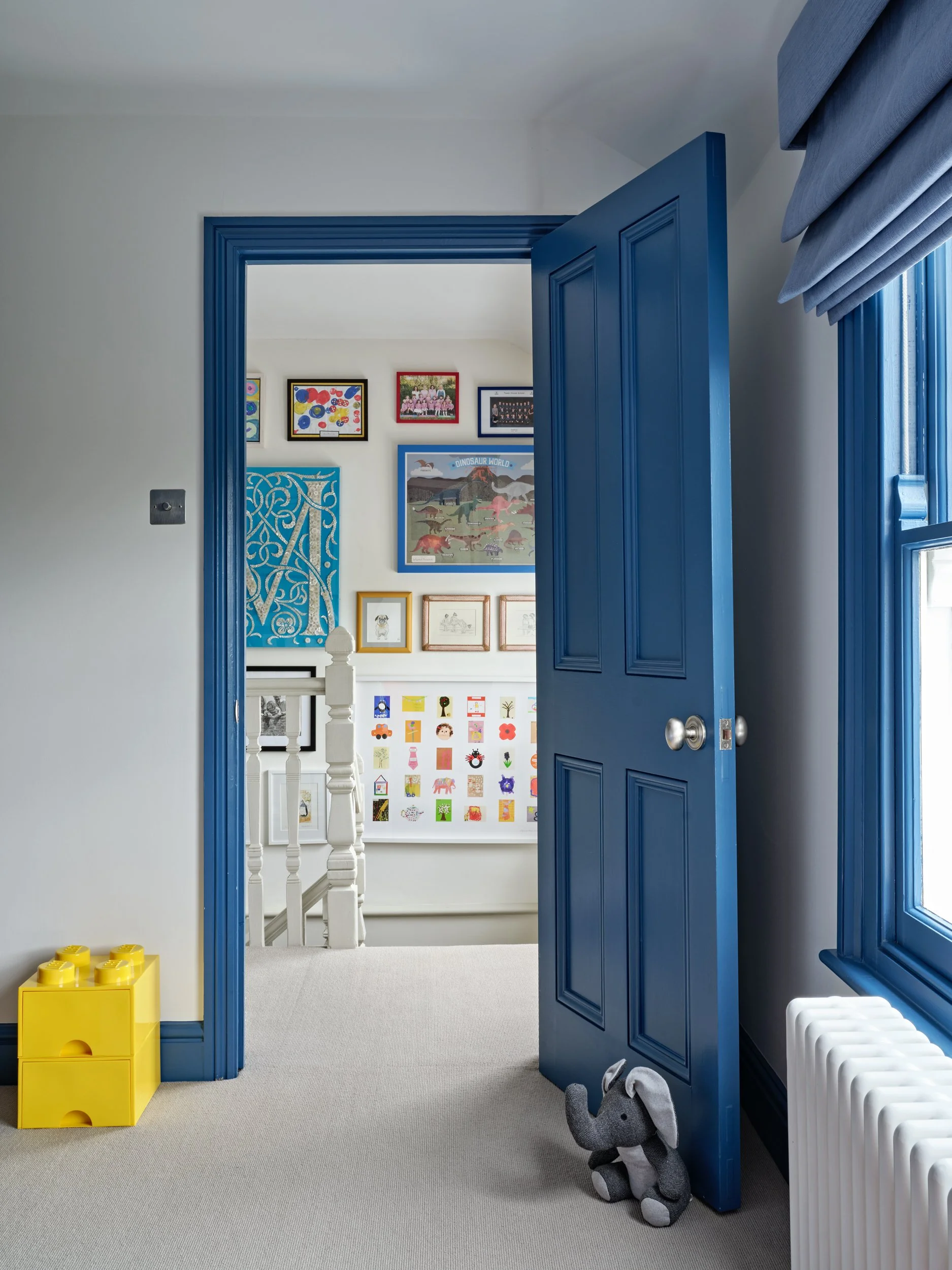 View of a house interior with a blue door open to a gallery wall with framed children's art and posters, a yellow storage cube, and a gray and white stuffed animal on the floor.