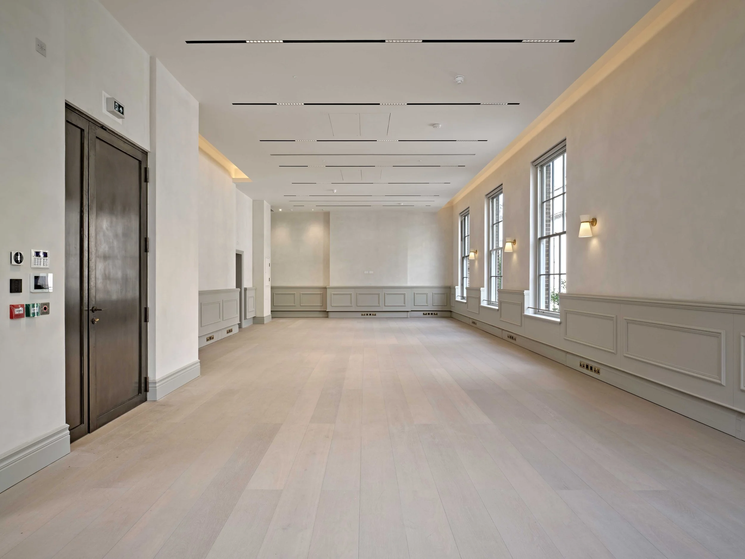 Empty, elegant hallway with large windows, white walls, decorative molding, and wall sconces lighting.