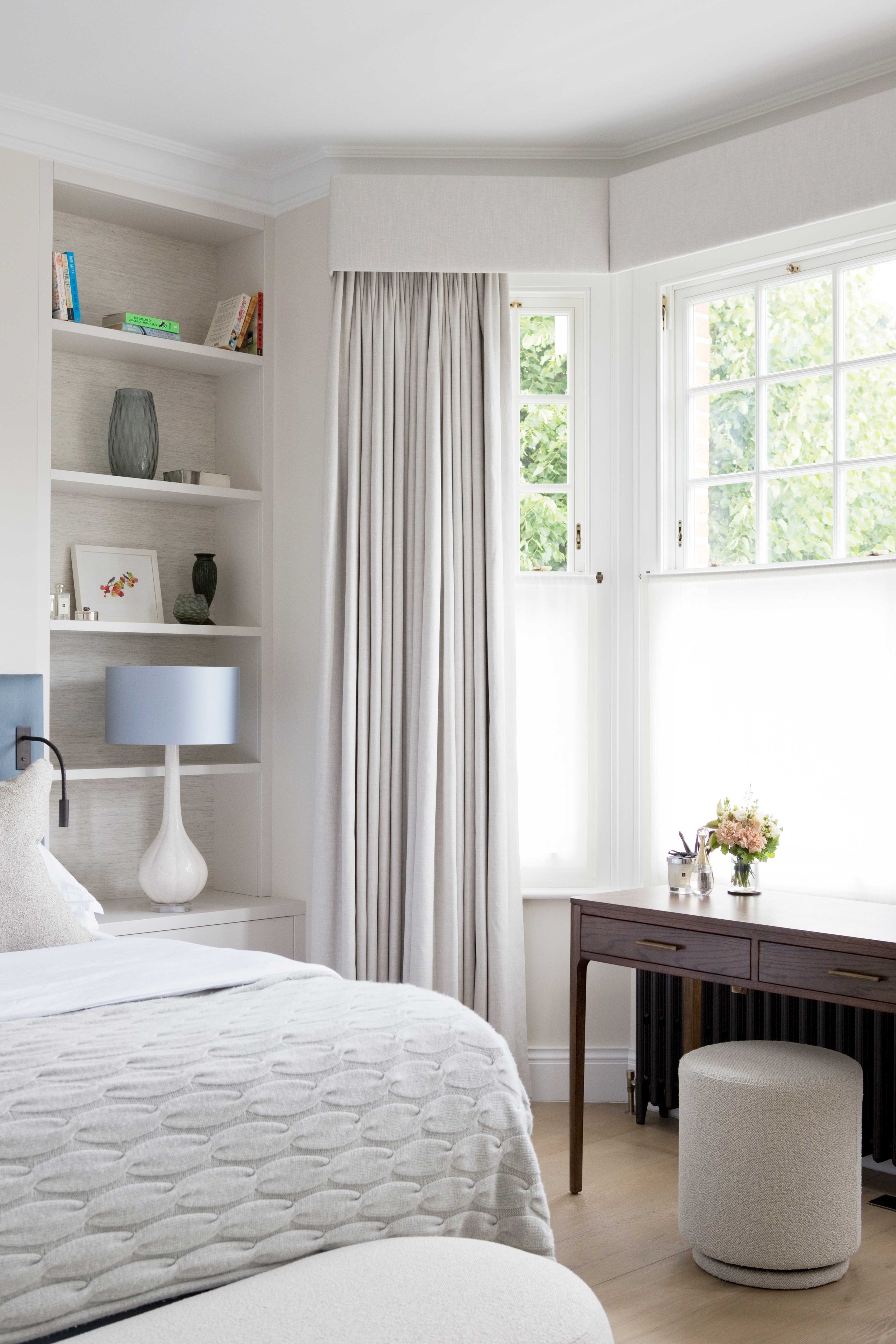 A cozy bedroom with a bed covered in a white textured quilt, a white bedside table with a white modern lamp, white built-in shelves with books and decorative items, a large window with sheer curtains and heavier drapes, a wooden console table with a 