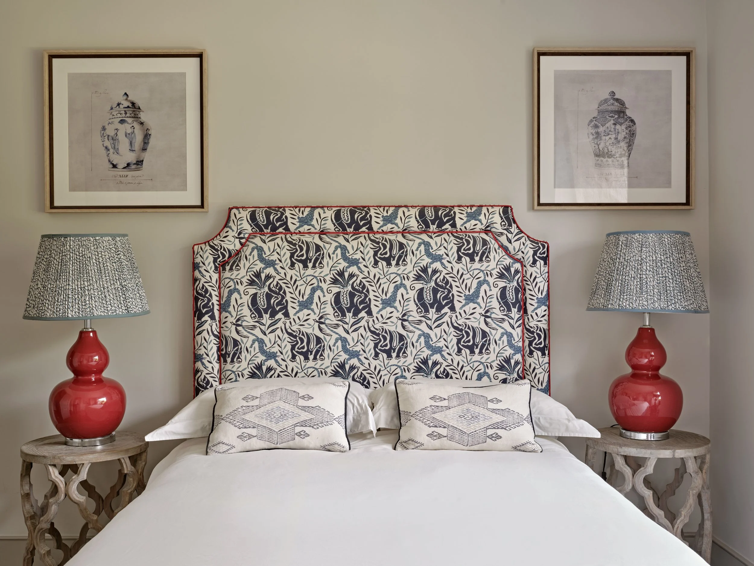A bedroom with a patterned headboard featuring elephants and foliage, two matching lamps with patterned shades on wooden side tables, a bed with white linens and decorative pillows, and framed vintage-style prints of vases hanging on a plain wall.