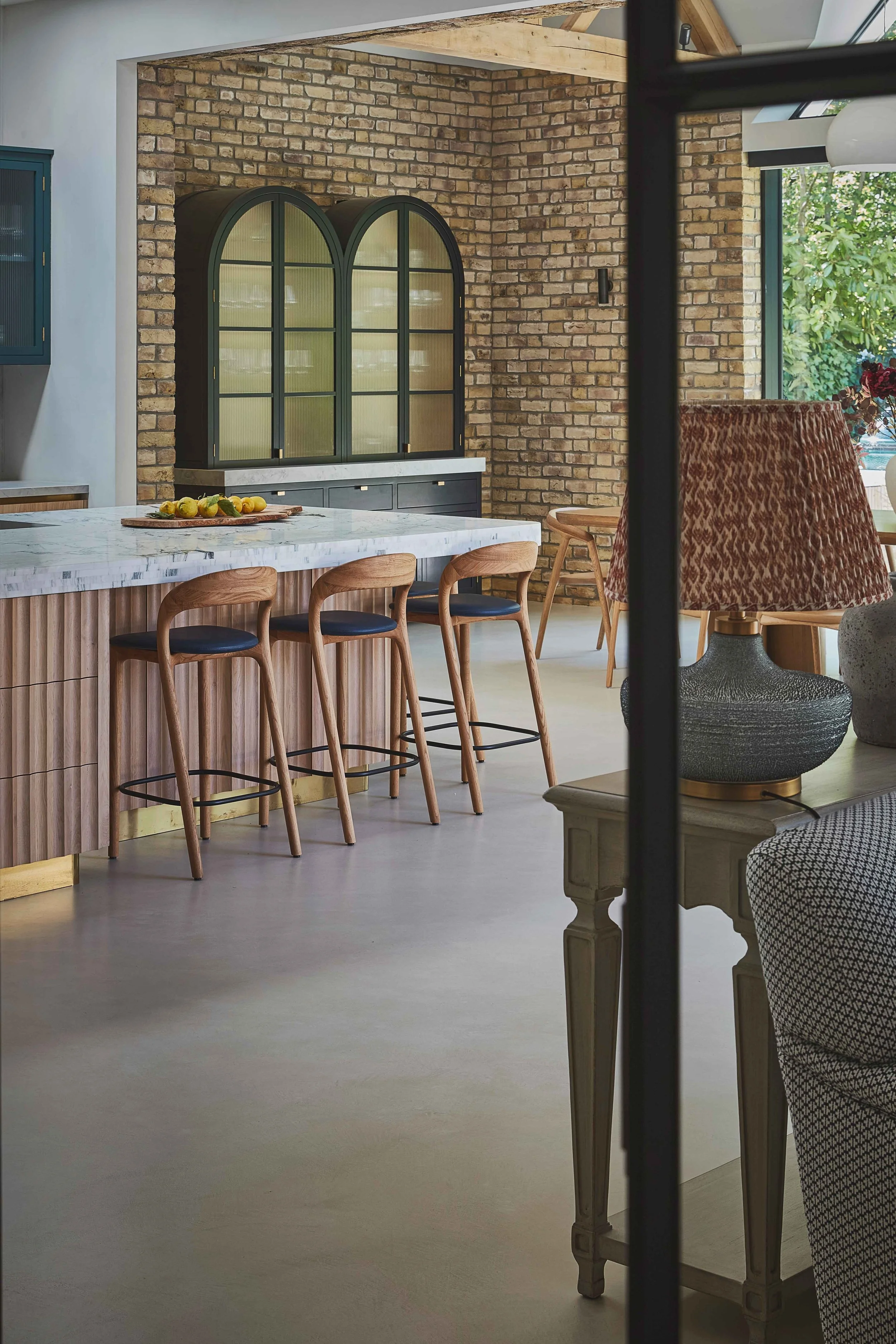 A modern kitchen with a white marble island counter, four wooden bar stools with dark blue seats, a brick wall background, and a hutch with arched glass doors. Part of the dining area is visible with a patterned upholstered chair and a table with a l