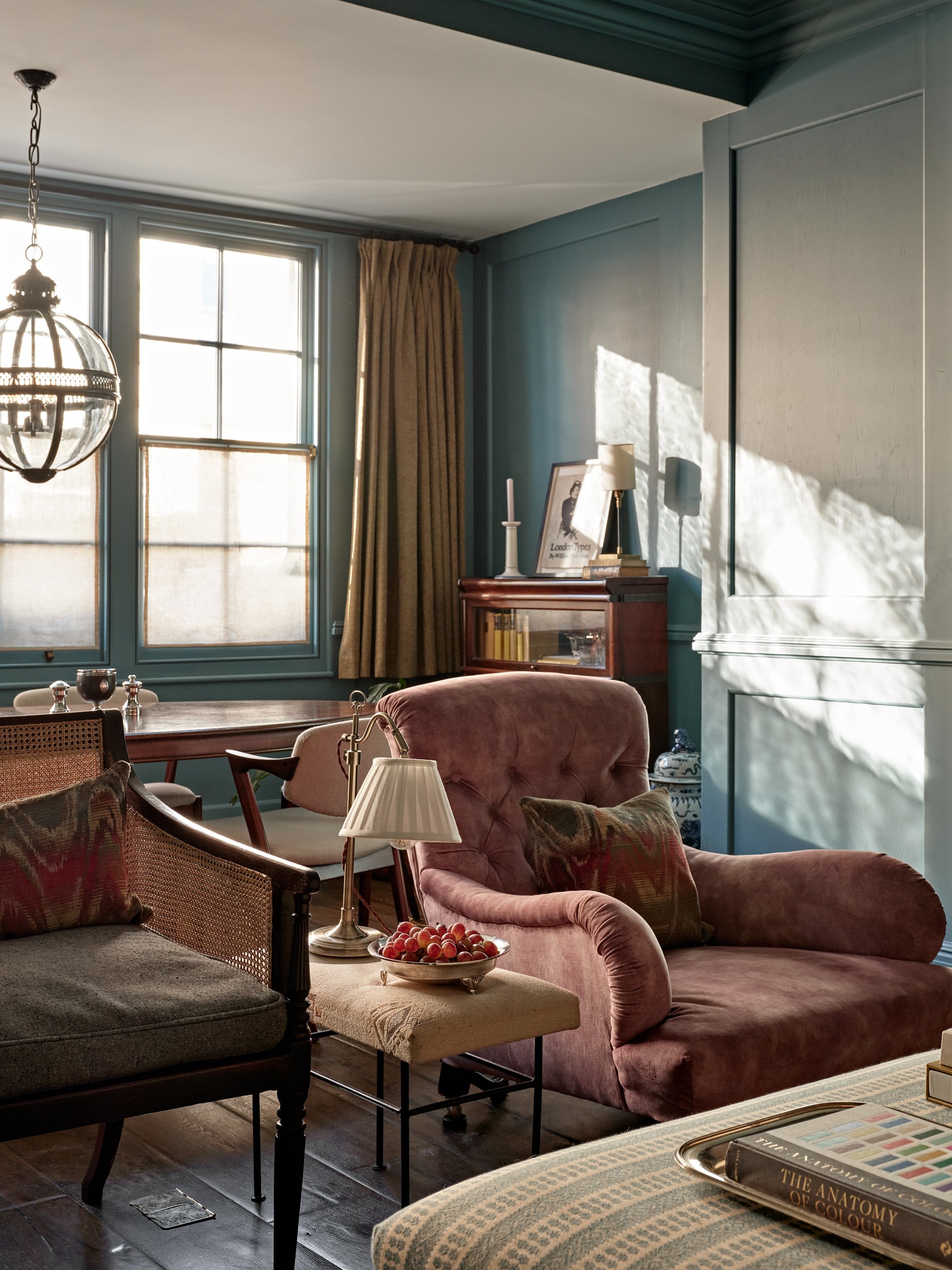 A cozy living room with teal walls, a window with beige curtains, a pink velvet armchair, a table with a bowl of cherries, and a bookshelf with framed artwork and decor.
