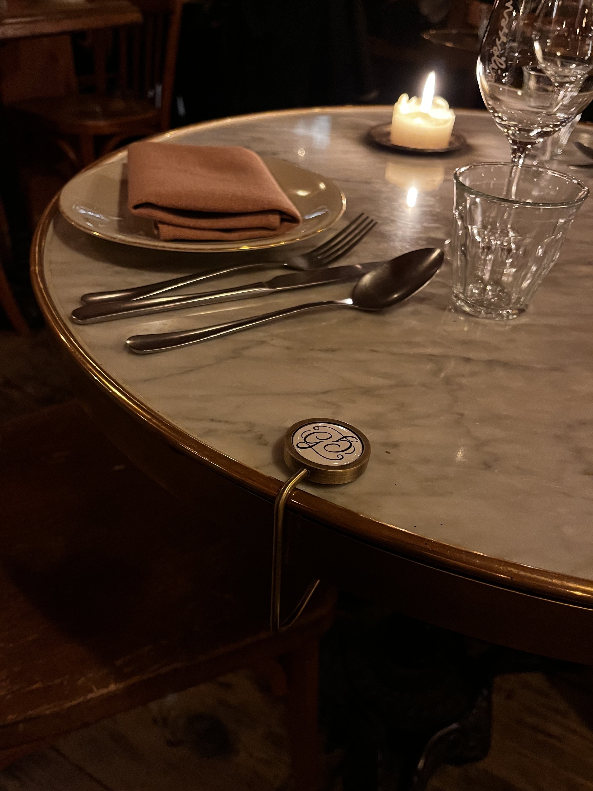 A marble table set for dining with a brown cloth napkin on a gold-rimmed plate, a fork, knife, and spoon arranged neatly, a lit candle, a  wine glass and a GANCIO bag hook.