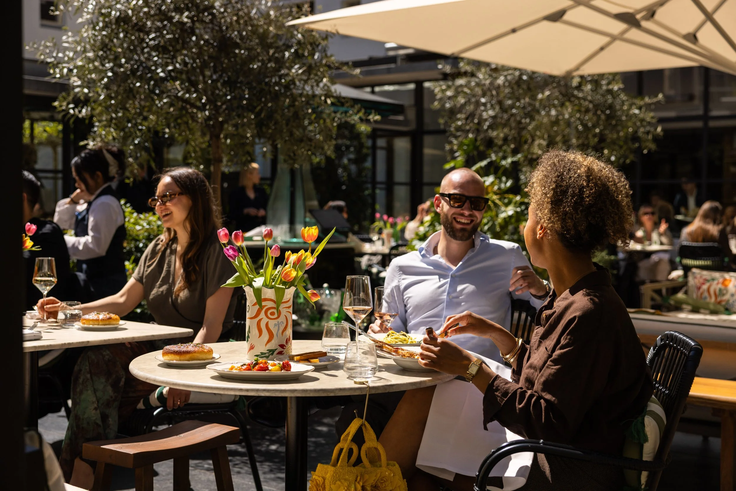People dining outdoors at a restaurant with a vase of flowers on the table, enjoying food and drinks during daytime.