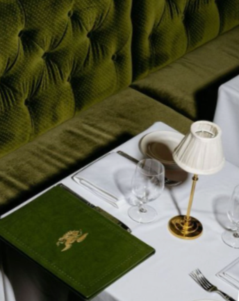 A neatly set dining table with a white tablecloth, wine glasses, a small table lamp, and a green menu or book. There is a green tufted upholstered seat in the background.