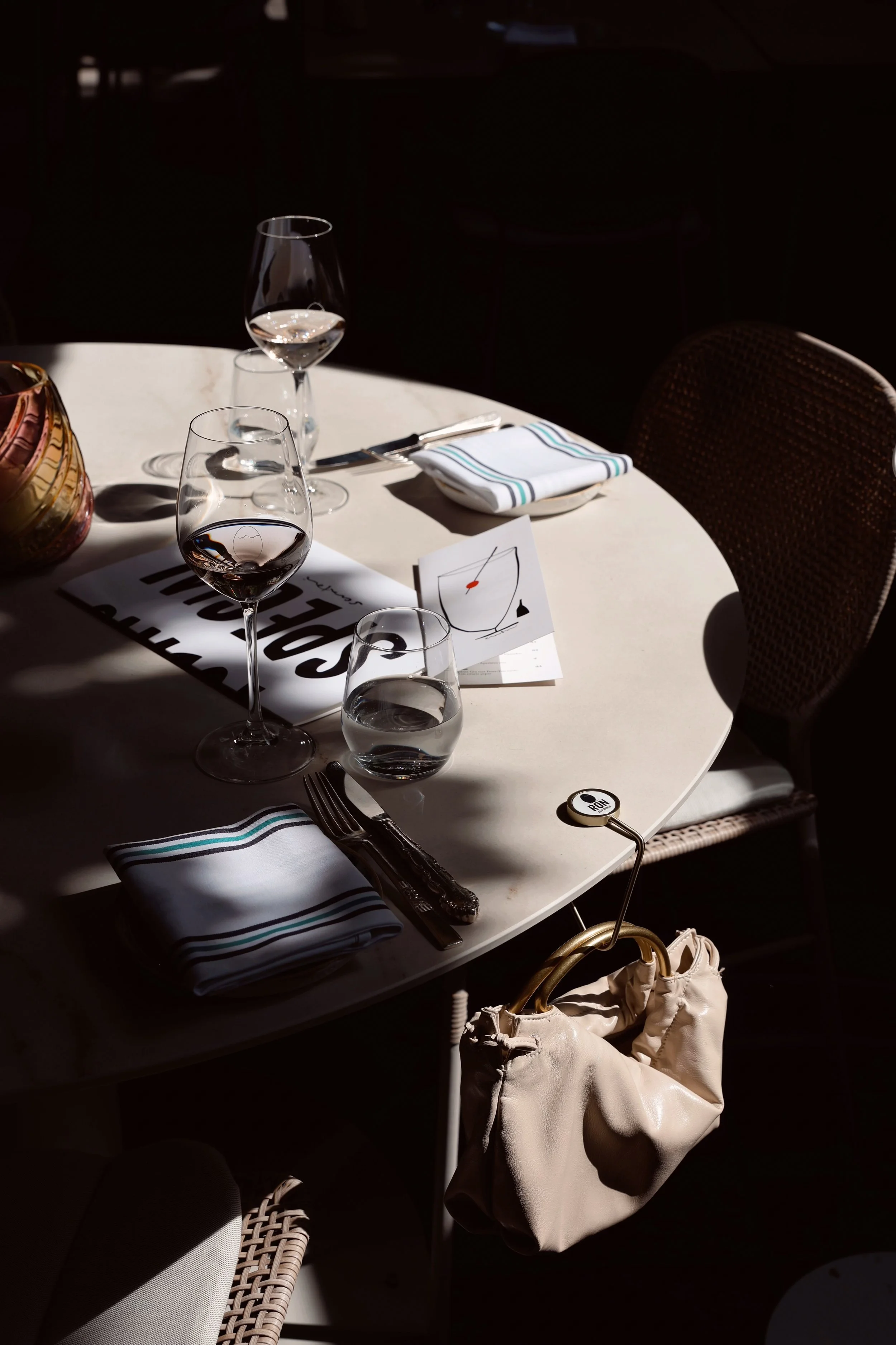 A round dining table set for a meal, with glasses of water and wine, napkins, cutlery, a small card with a simple line drawing of a glass and a GANCIO bag hook.