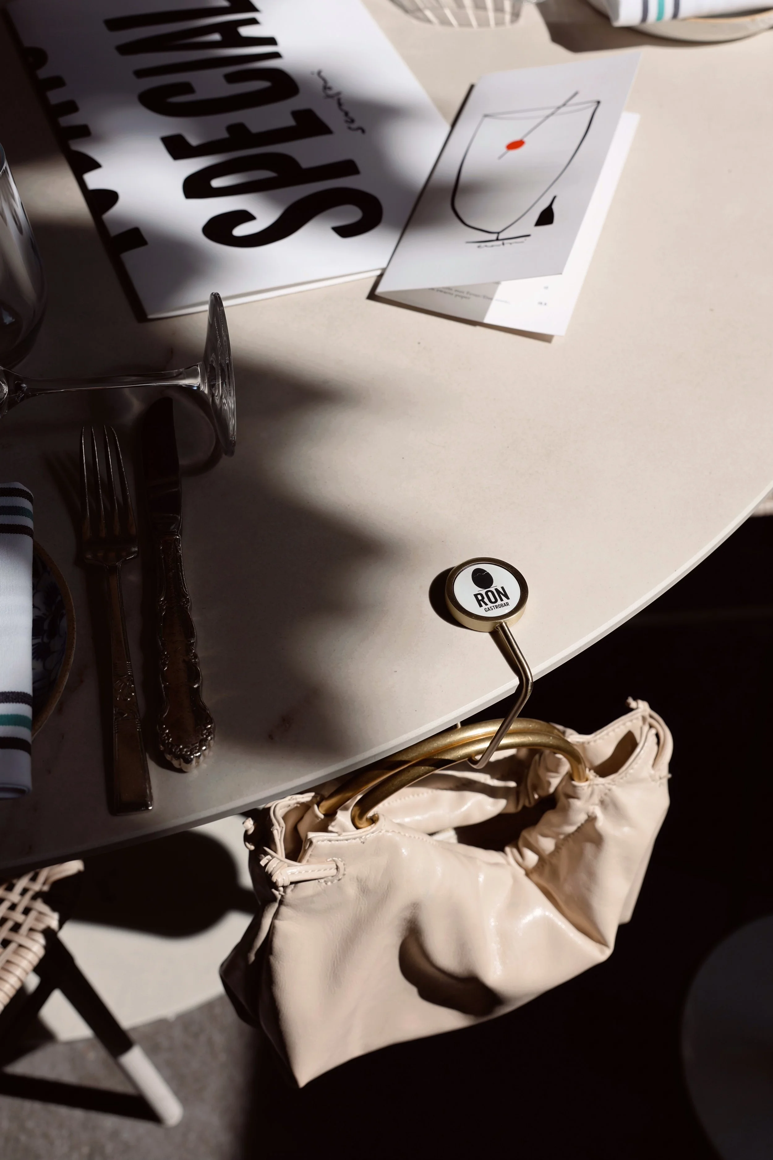 Part of a dining table with a wine glass, silverware, a white bag, art prints, and a customized GANCIO bag hook.