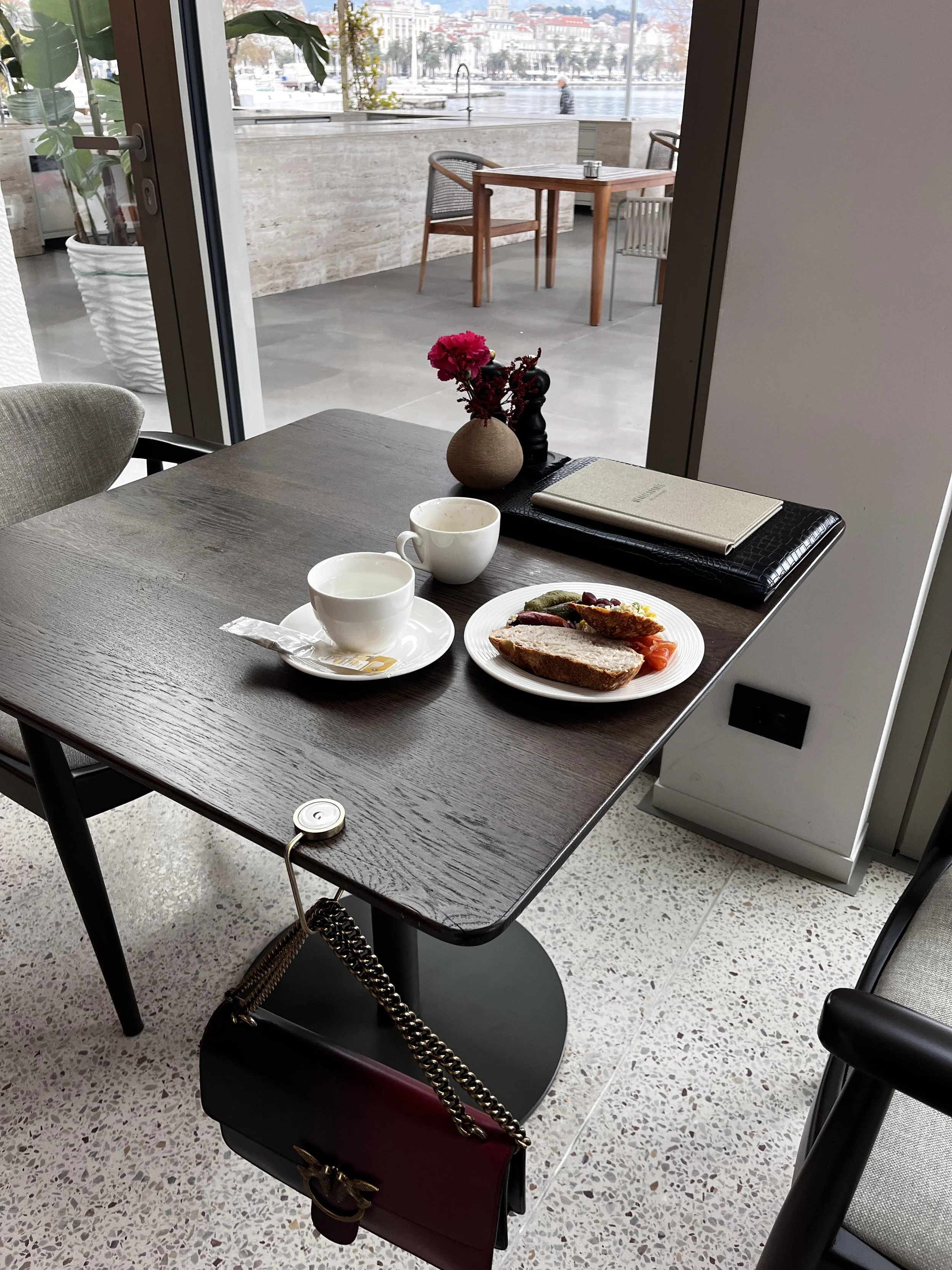 Interior of a cafe with a wooden table set for breakfast, including a plate of bread, a cup of coffee and a GANCIO bag hook. Outside there is an outdoor patio with tables and chairs overlooking a waterfront with boats and buildings.
