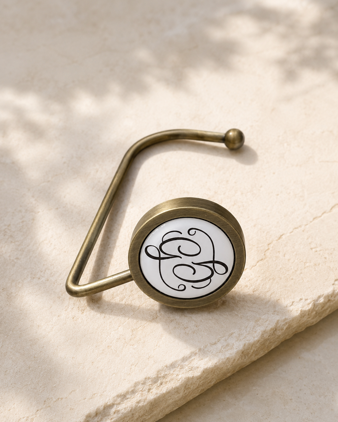 A brass bag hook with a featuring black cursive monogram initials 'G' on a white background, resting on a light-colored stone surface.