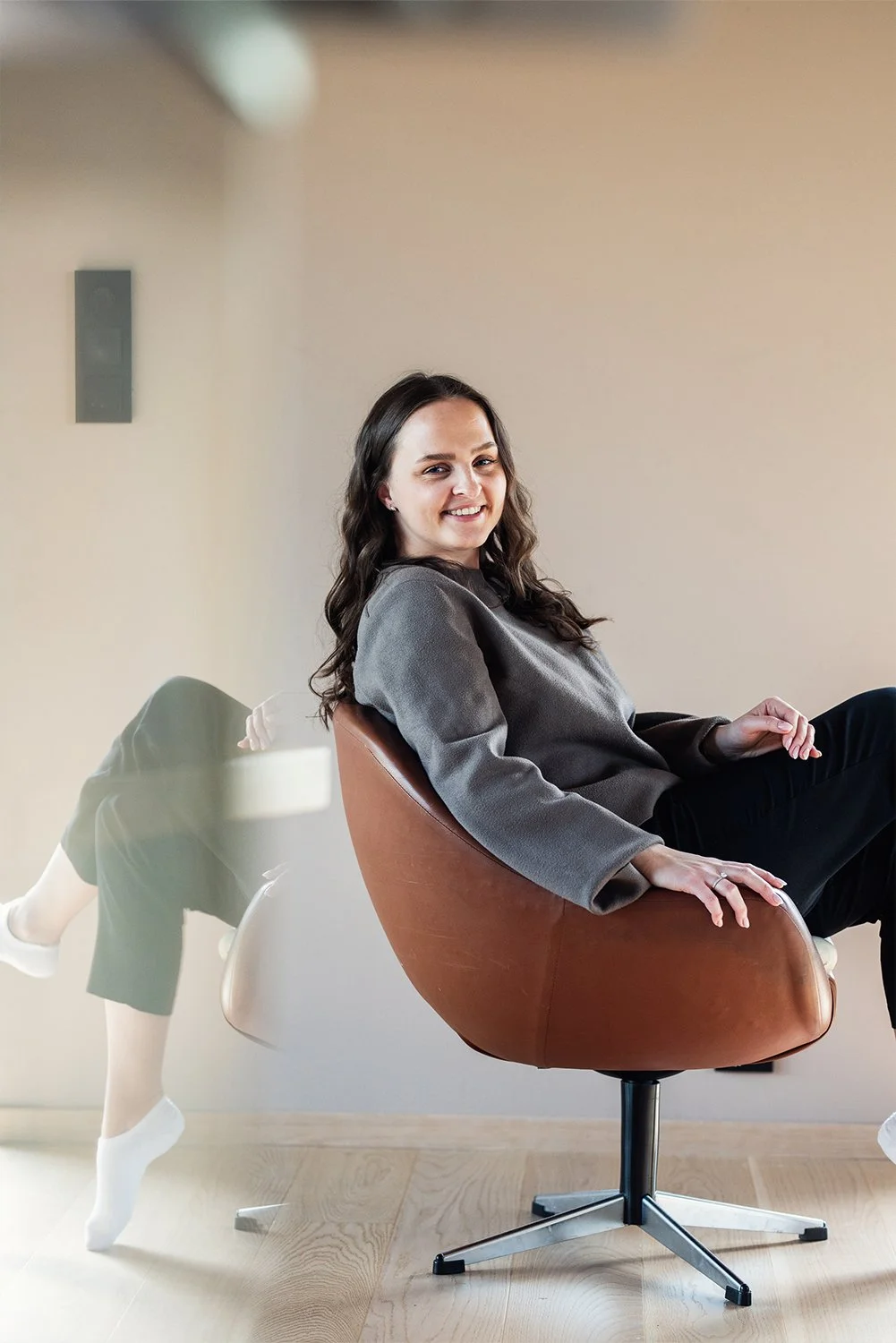 En kvinne sitter i en moderne stol, smilende mot kamera, i et rom med lys vegg og tregulv. En refleksjon av en annen person som står eller ligger er synlig i masken på stolens side.