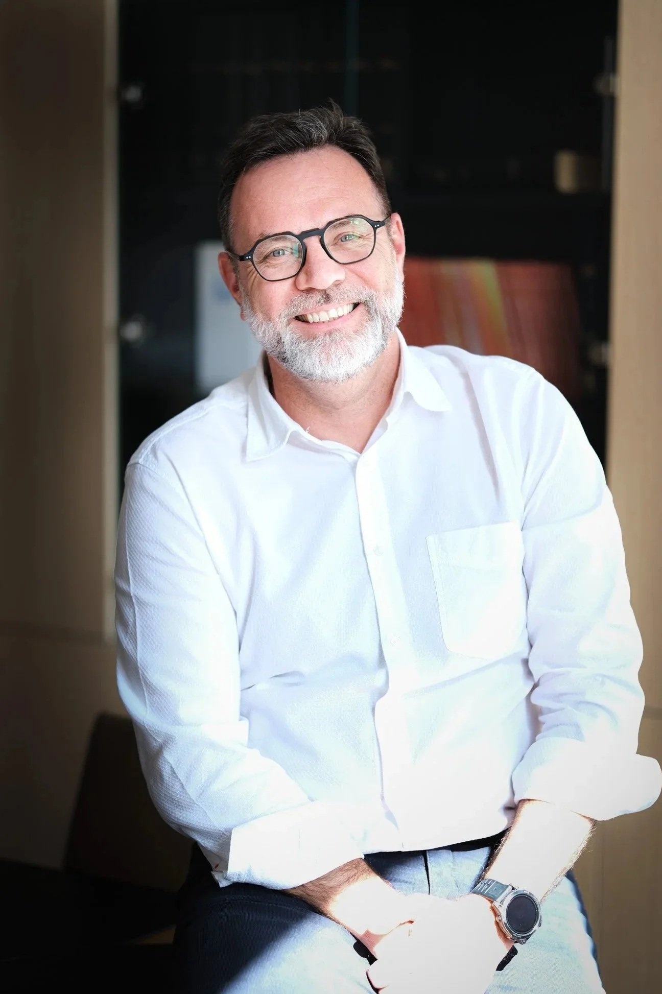 Un homme souriant avec des lunettes rondes, une barbe grise, portant une chemise blanche et une montre, assis dans un intérieur moderne.
