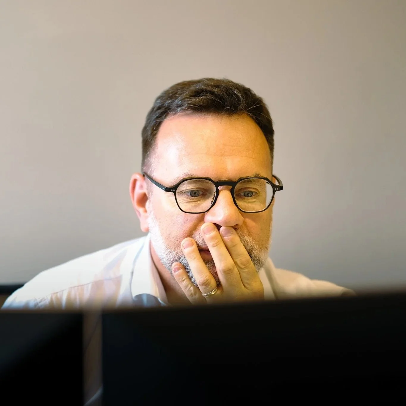 Homme portant des lunettes, ayant une barbe, en train de regarder un écran d'ordinateur, la main sur le visage, dans un environnement de bureau.