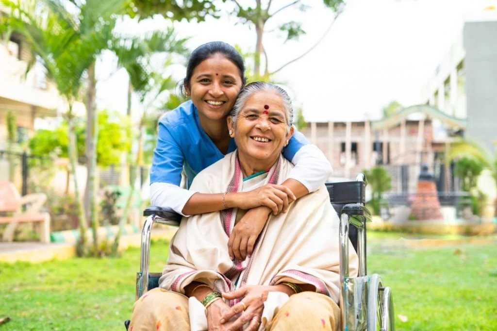 A young woman in a blue uniform hugging an elderly woman in a wheelchair outdoors.