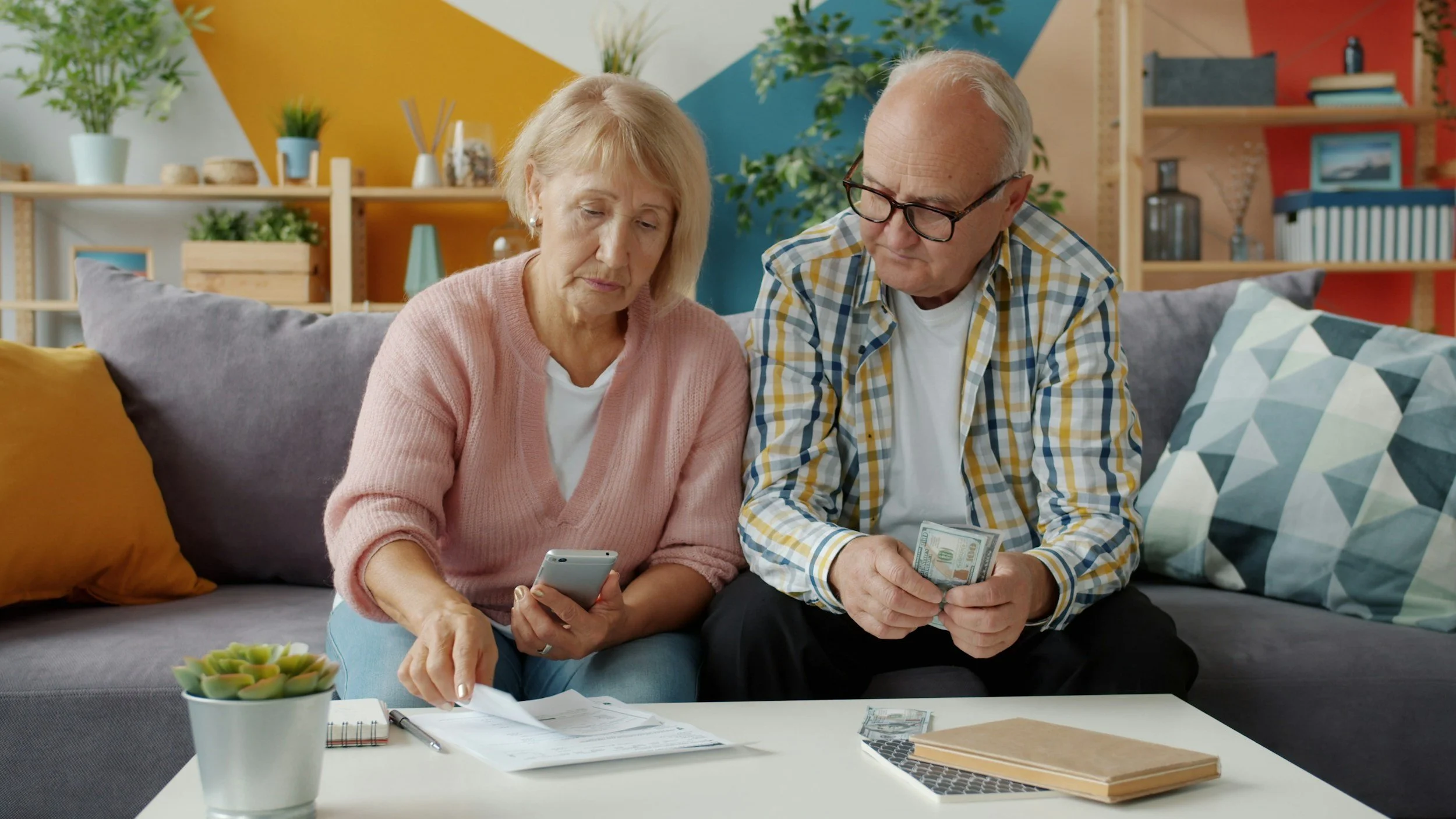 An elderly woman and man sitting on a couch, looking at a smartphone and holding a stack of money, with financial papers and notebooks on the table in front of them.