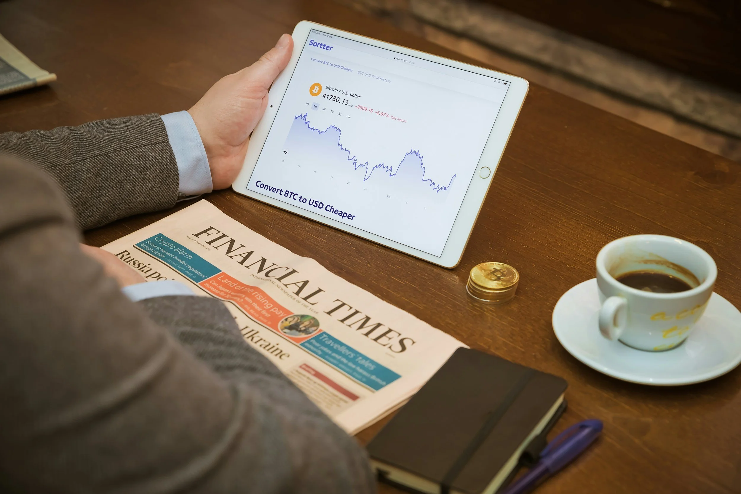 Person holding a tablet displaying a Bitcoin price chart with a London Times newspaper, a cup of coffee, and stacks of gold coins on a wooden table.