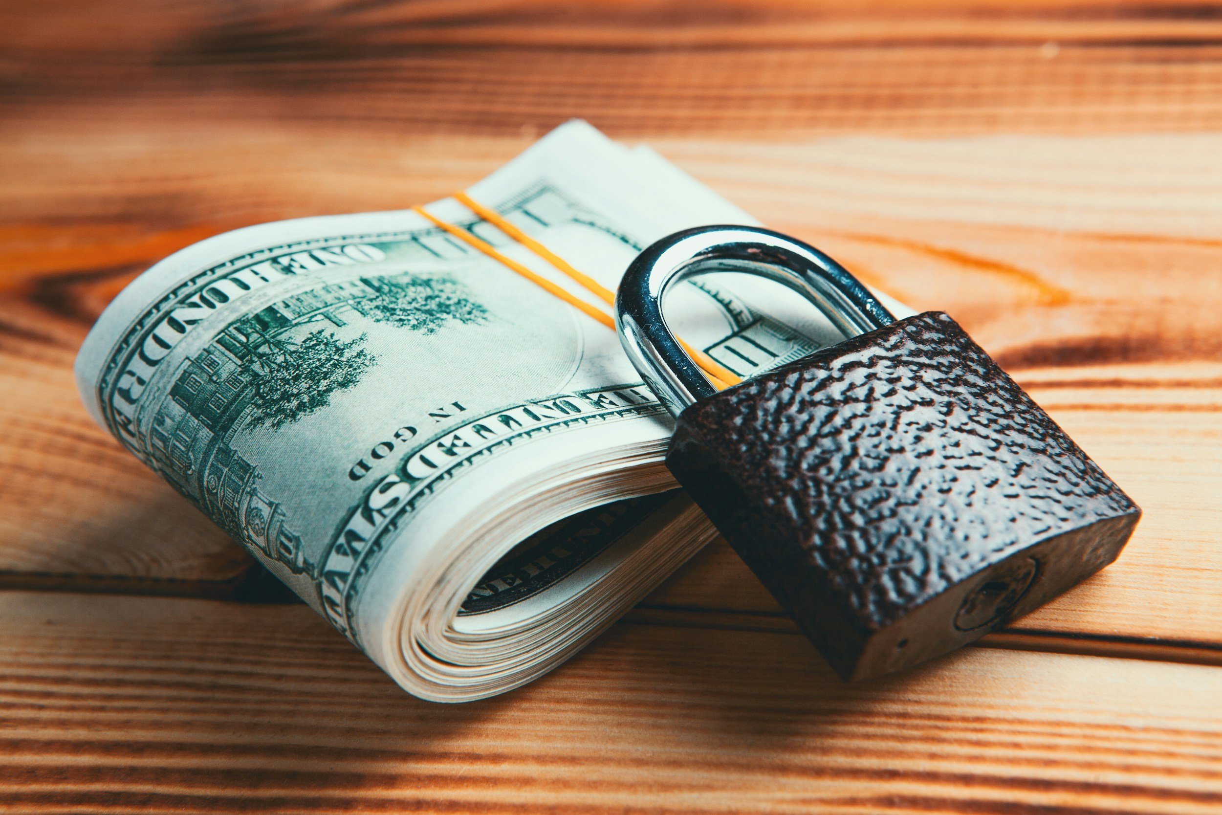 A rolled bundle of U.S. hundred-dollar bills secured with an orange rubber band, placed on a wooden surface with a textured padlock fastening the bundle.