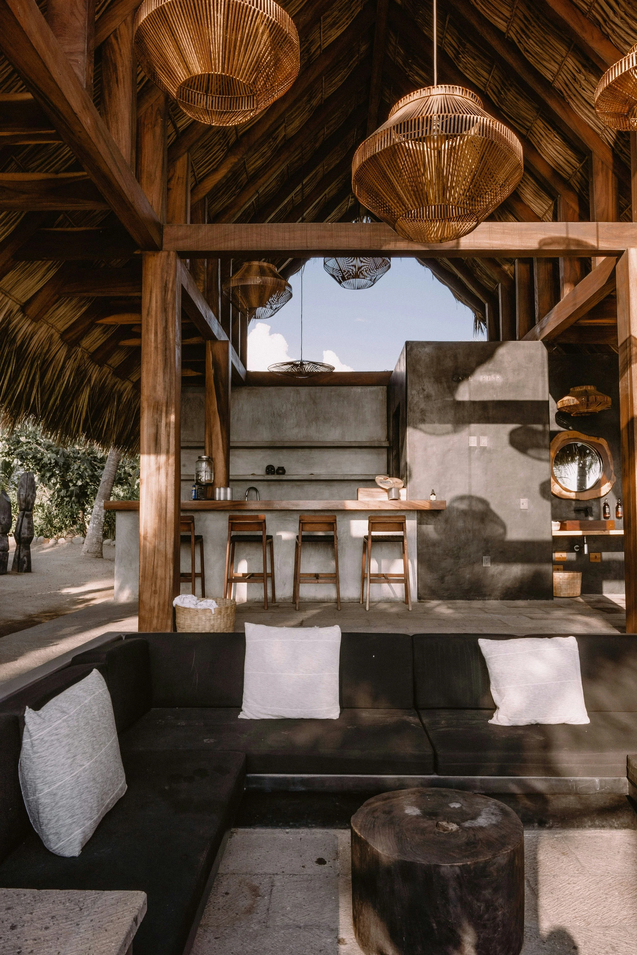 Open-air tropical bar with wooden structure, thatched roof, wicker chandeliers, concrete walls, a bar counter with stools, and dark cushioned seating with white pillows.