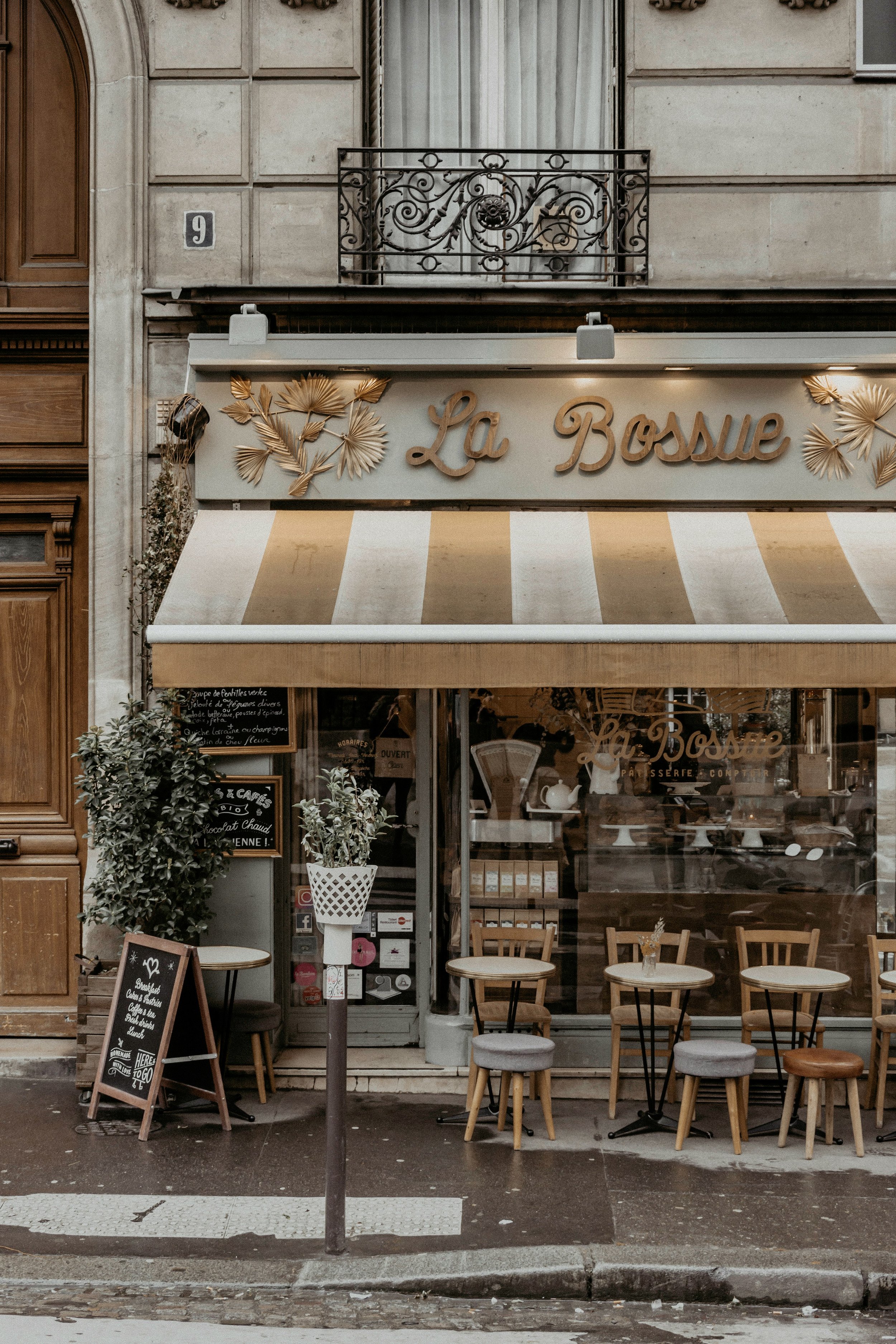 Exterior view of a café named La Bossie with a striped beige and white awning, wooden chairs and tables outside, small potted plants, and handwritten chalkboard menus near the entrance. The building has a stone facade with a small decorative wrought iron balcony above.