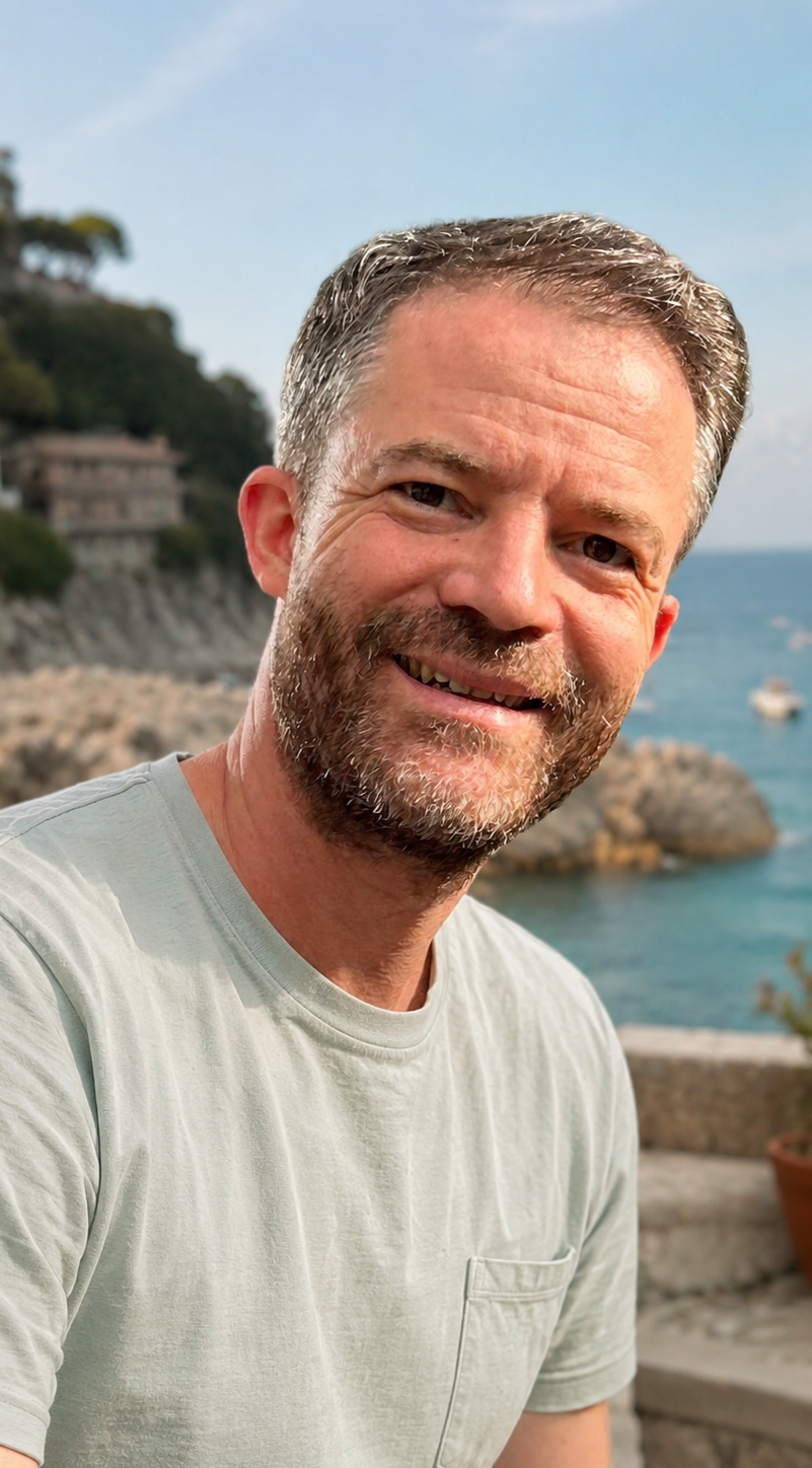 A smiling middle-aged man with short hair and a beard, outdoors near the coast with rocks, trees, and a boat in the background.