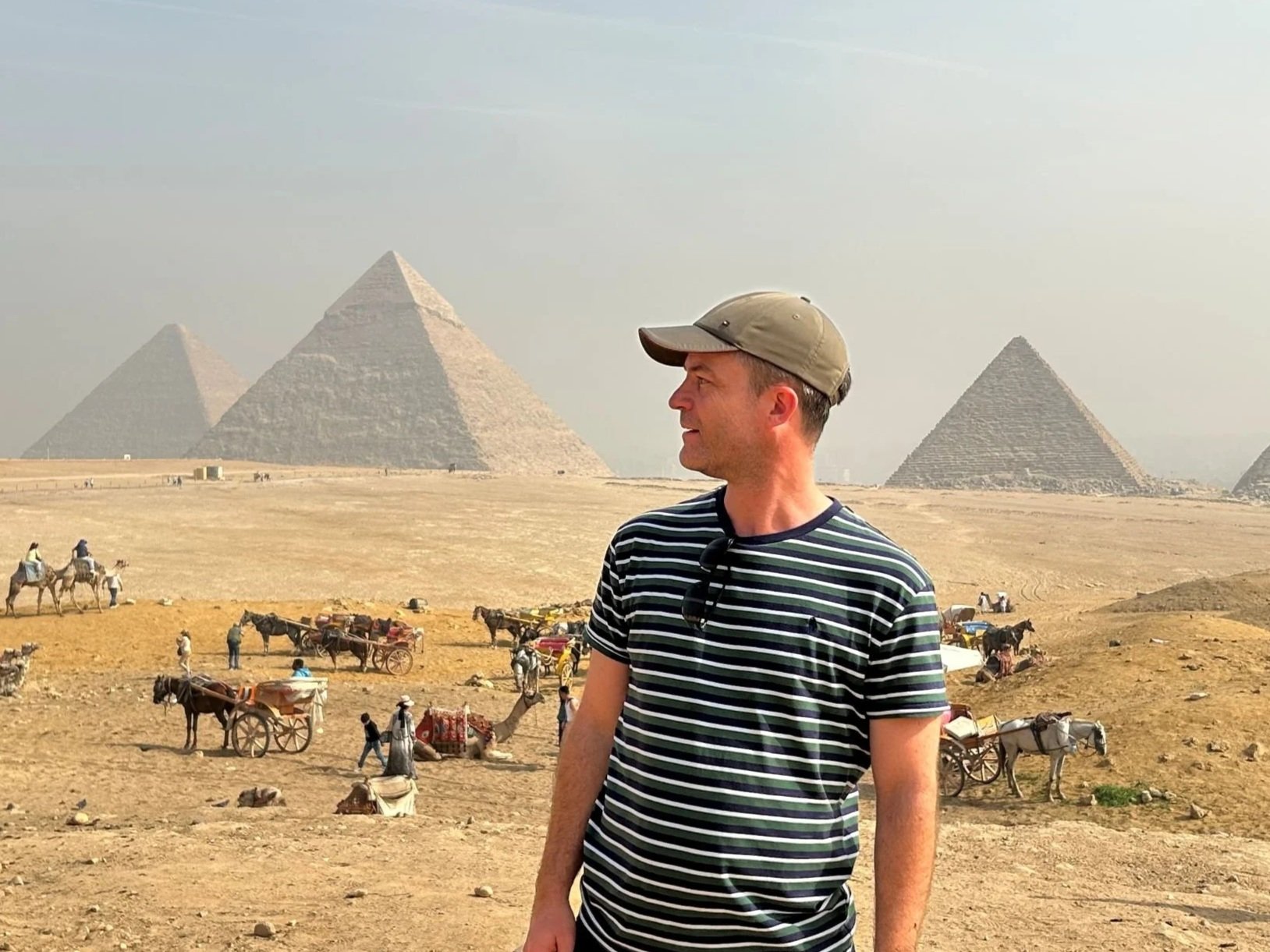 A man wearing a striped shirt and a khaki cap standing in a desert landscape with the pyramids of Egypt in the background. There are camels and horse-drawn carts with people in the distance.