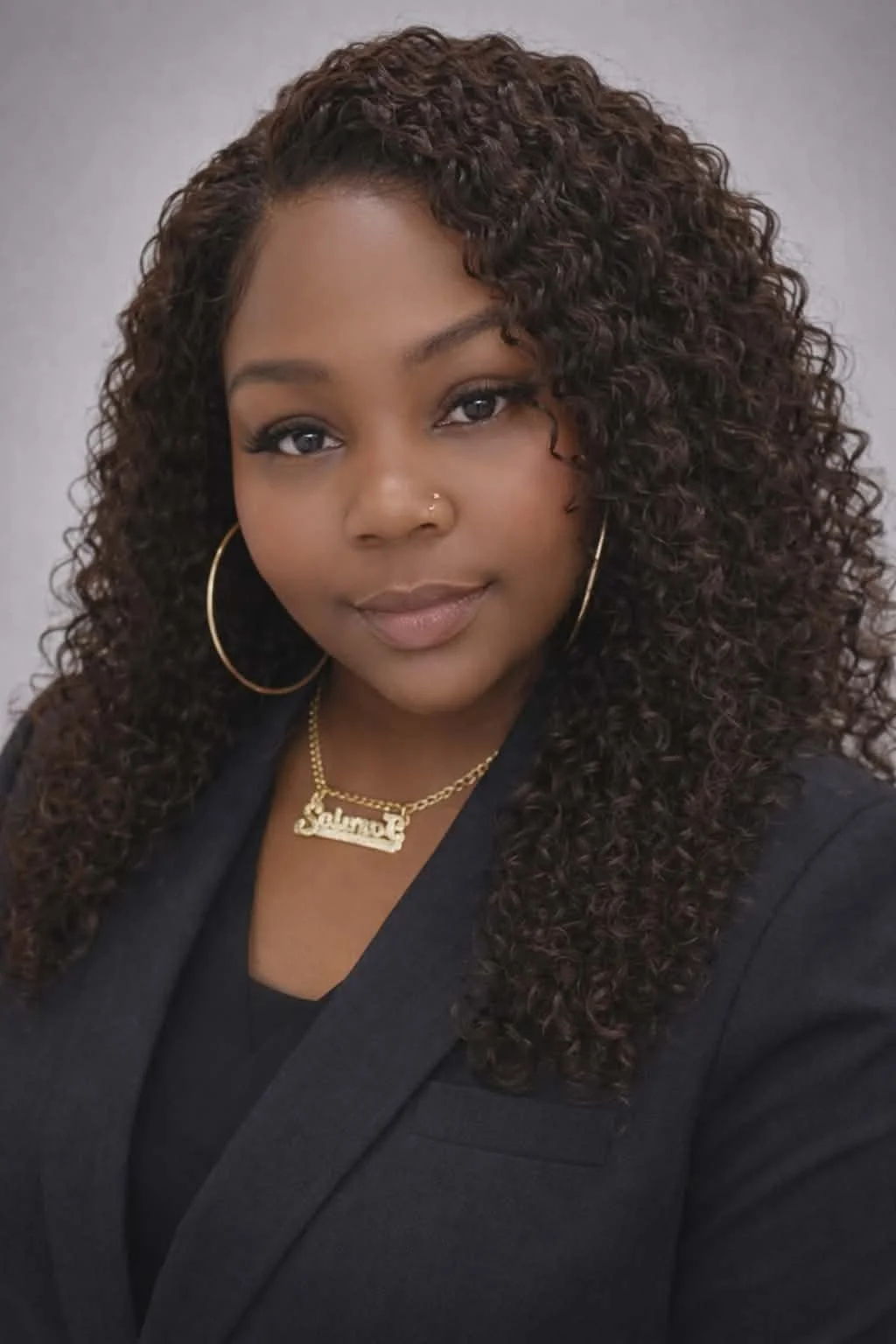 A woman with curly dark brown hair, wearing gold hoop earrings and a gold necklace with a nameplate, dressed in a black blazer, looking at the camera with a slight smile, against a neutral background.