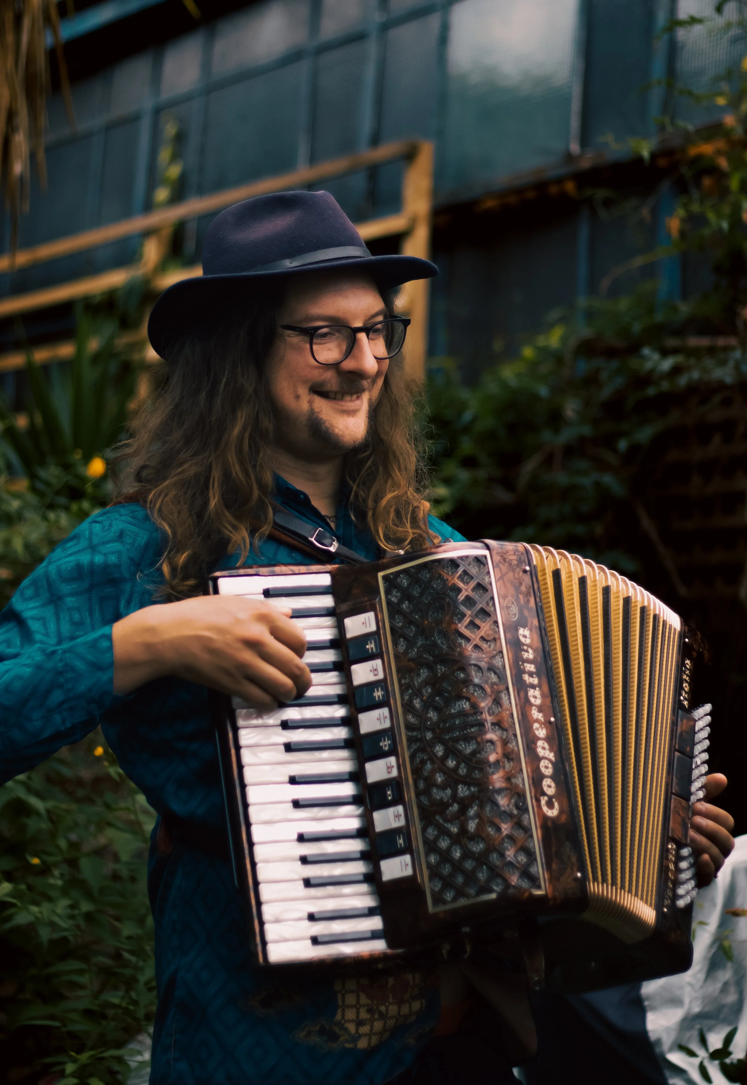 Person wearing glasses and a dark hat playing an accordion outdoors, surrounded by greenery.