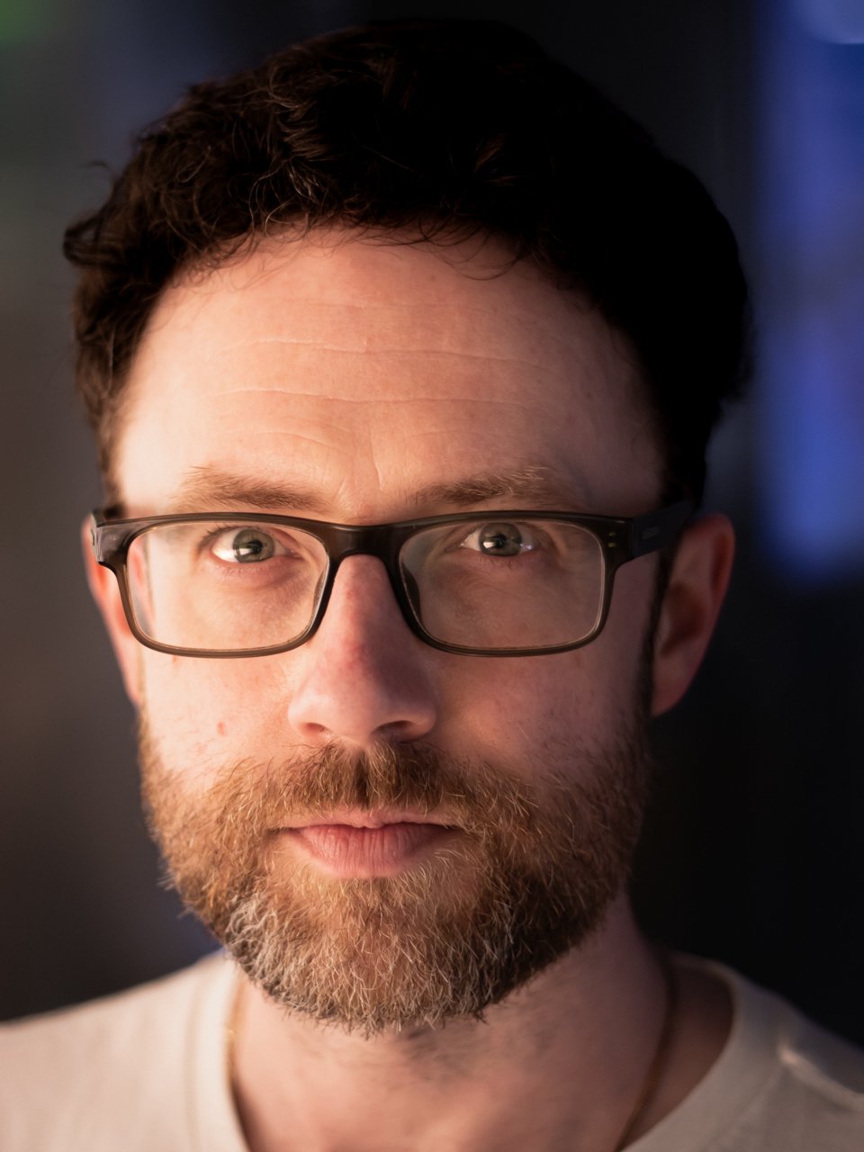 Close-up of a man with glasses and a beard, looking directly at the camera against a dark background.