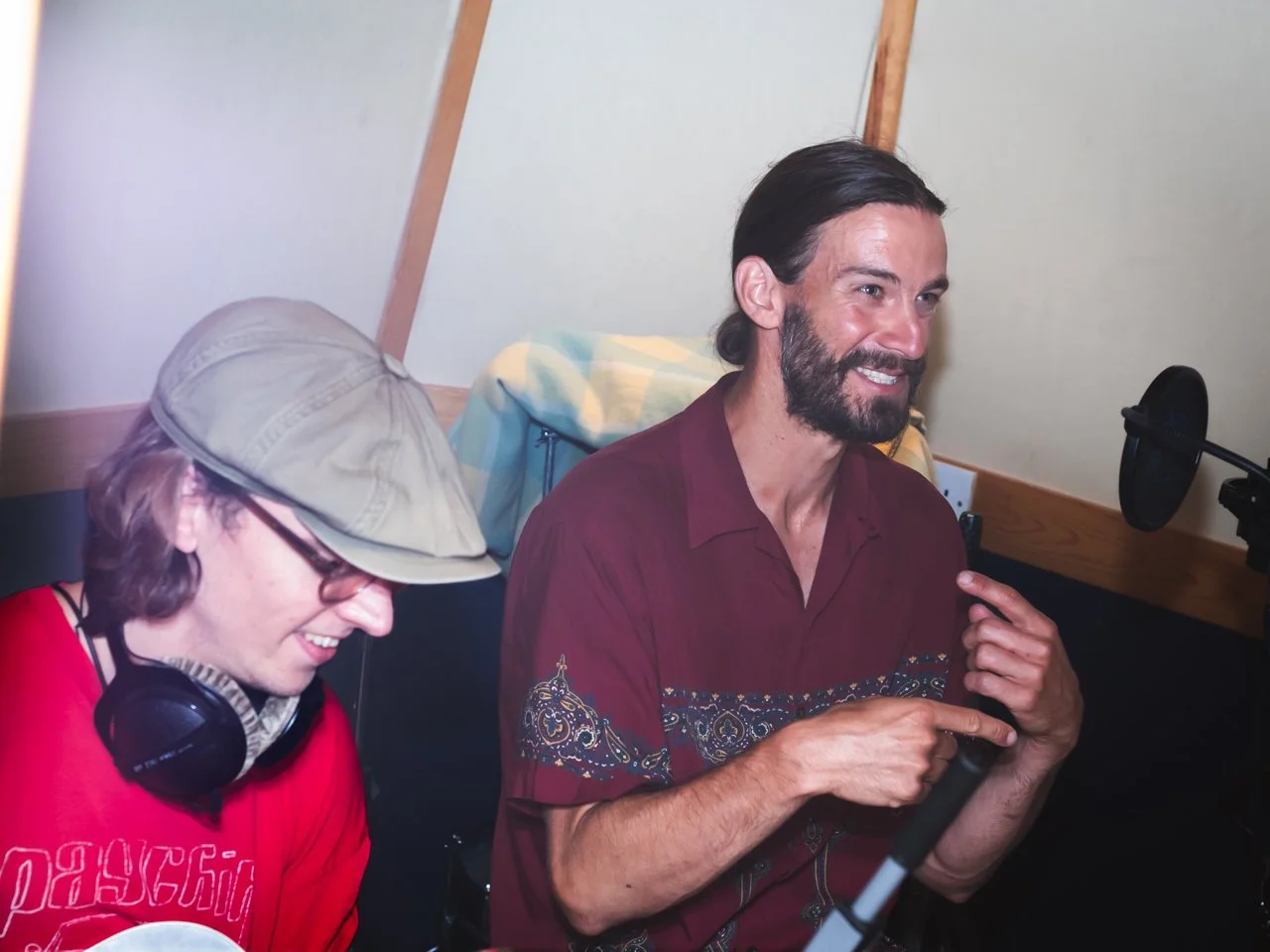 Two men in a recording studio, one wearing a beige cap and headphones, smiling, the other in a maroon shirt with a beard, pointing at the microphone.
