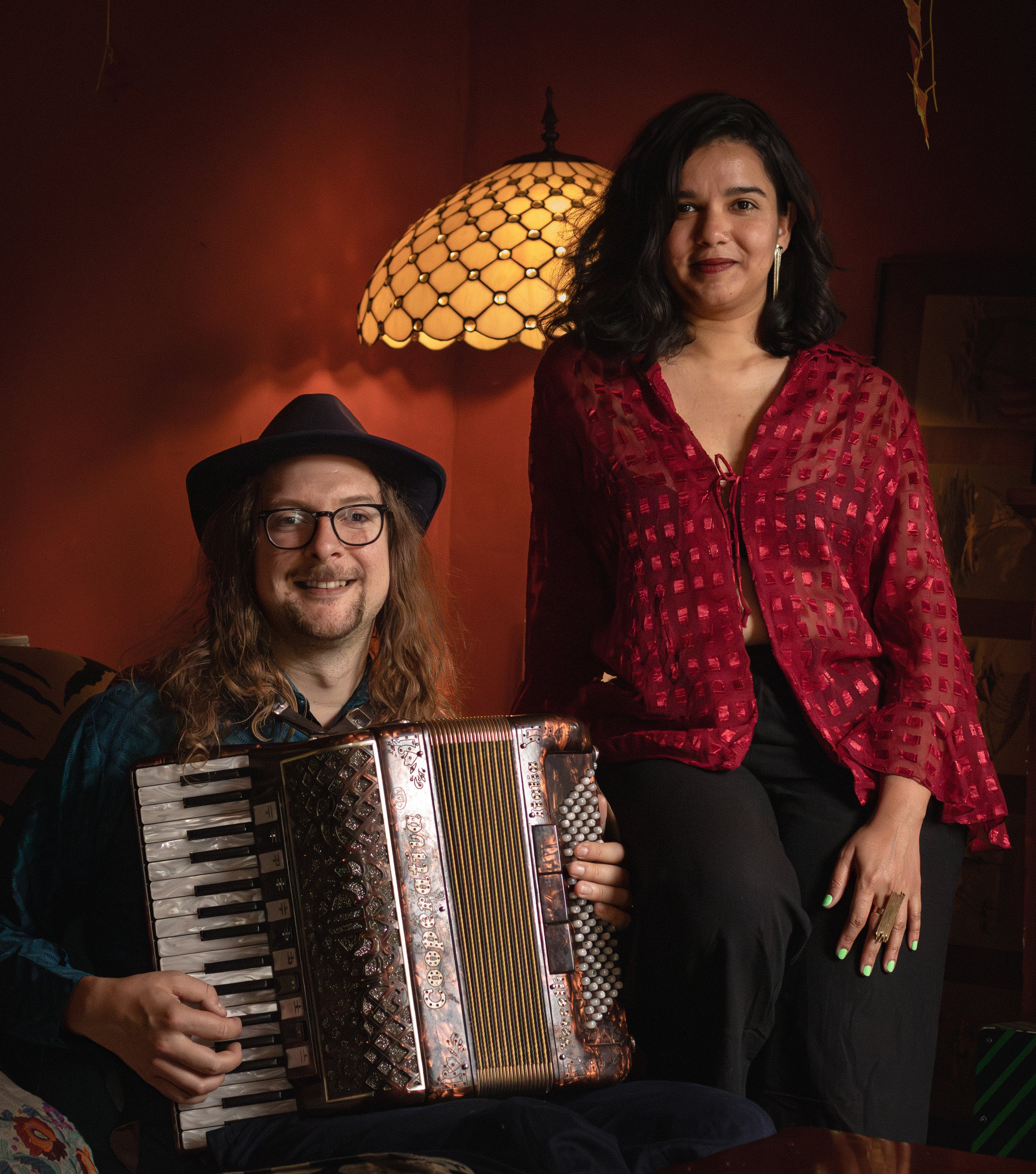Two women in a dimly lit room, one playing an accordion, the other standing nearby, with a warm, amber-colored lamp and wall decorations in the background.