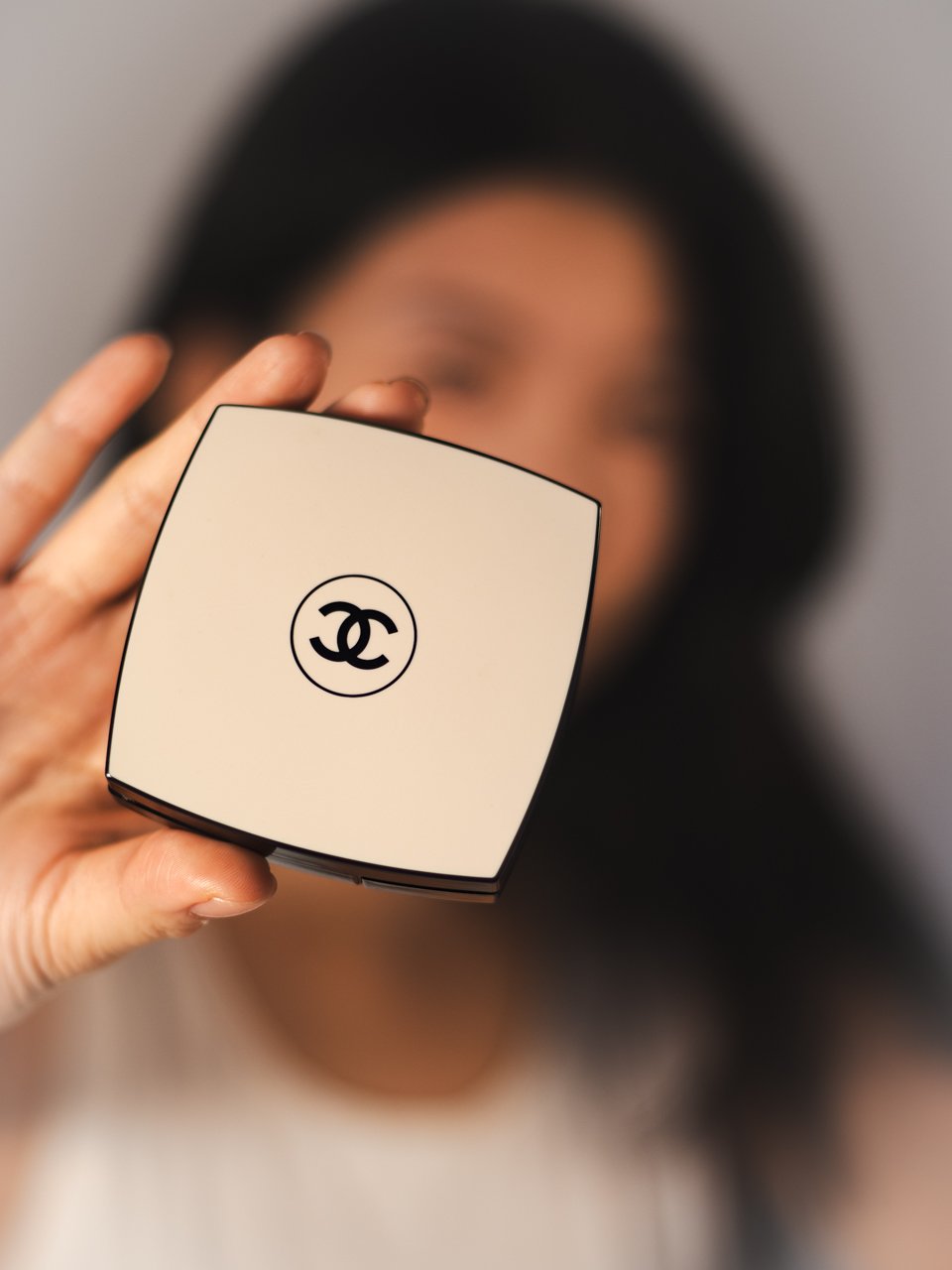 Person holding a white compact with the Chanel logo, blurred face in the background.
