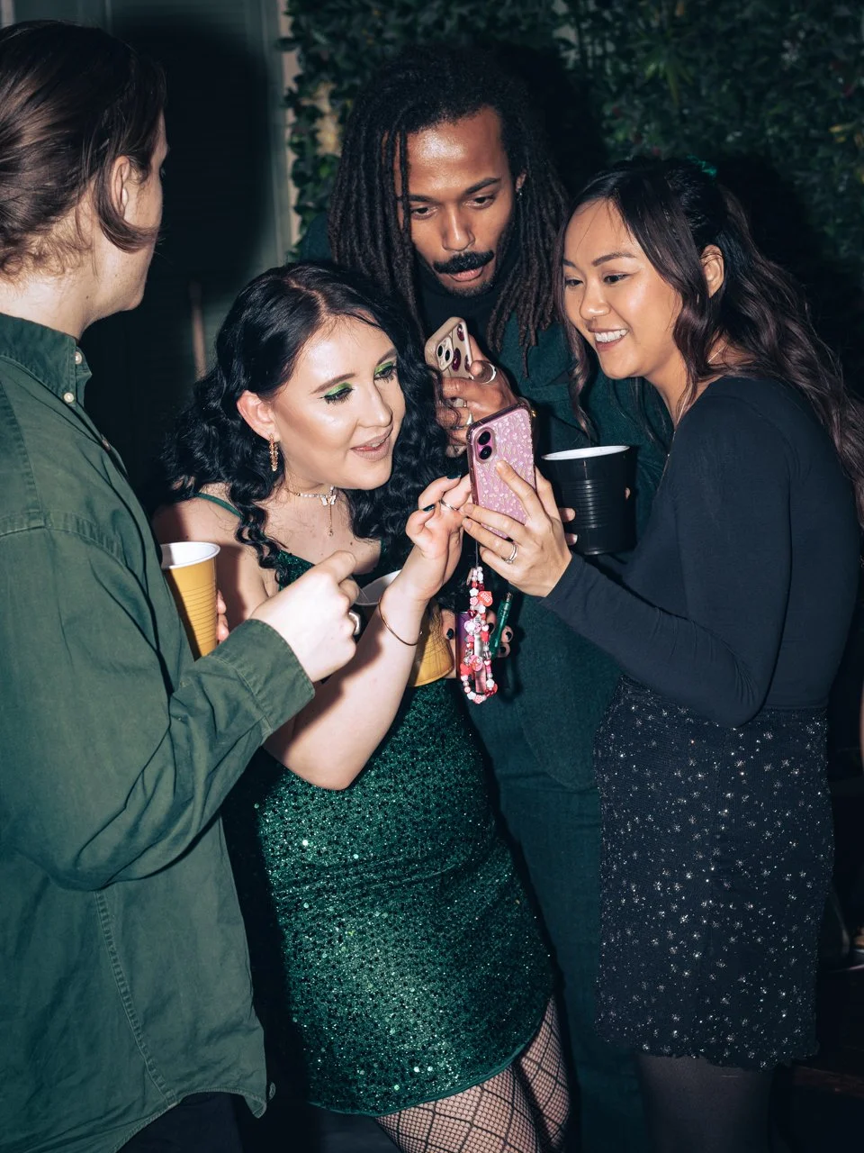 Four friends at a party looking at a phone together, with drinks in hand, wearing festive clothing.