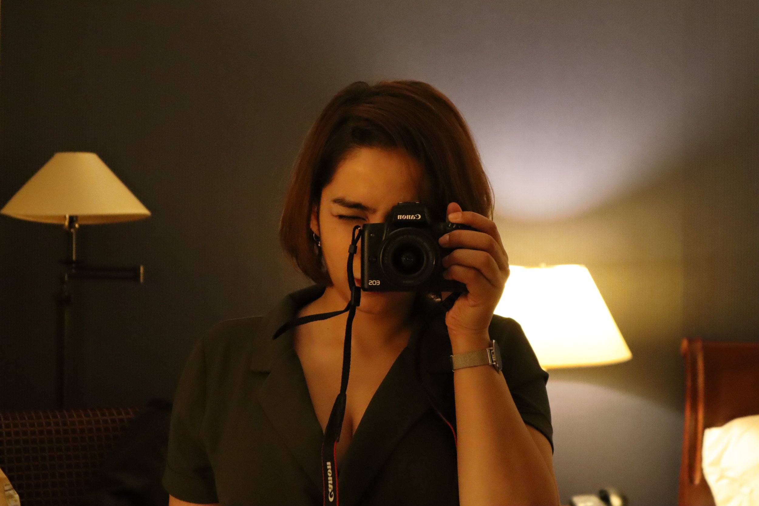 A person with short dark hair holding a camera takes a mirror selfie in a dimly lit room with a beige lamp and a dark wall background.