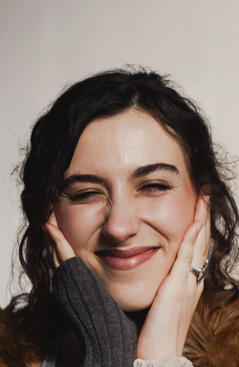Close-up of a woman smiling and scrunching her face, with hands on cheeks, wearing a dark gray sweater and a ring on her right hand.