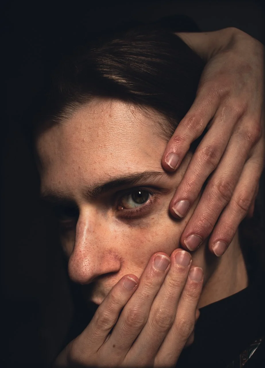 Close-up portrait of a person with light skin, dark hair, and green eyes, with their hands framing their face, taken in a dark room only illuminating his face for headshot.