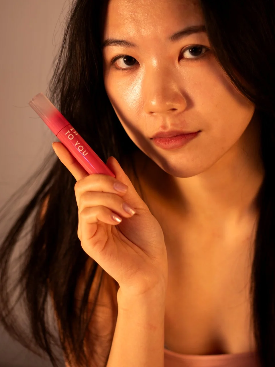 A young woman with long dark hair holding a pink tube of lipstick close to her face, looking directly at the camera.