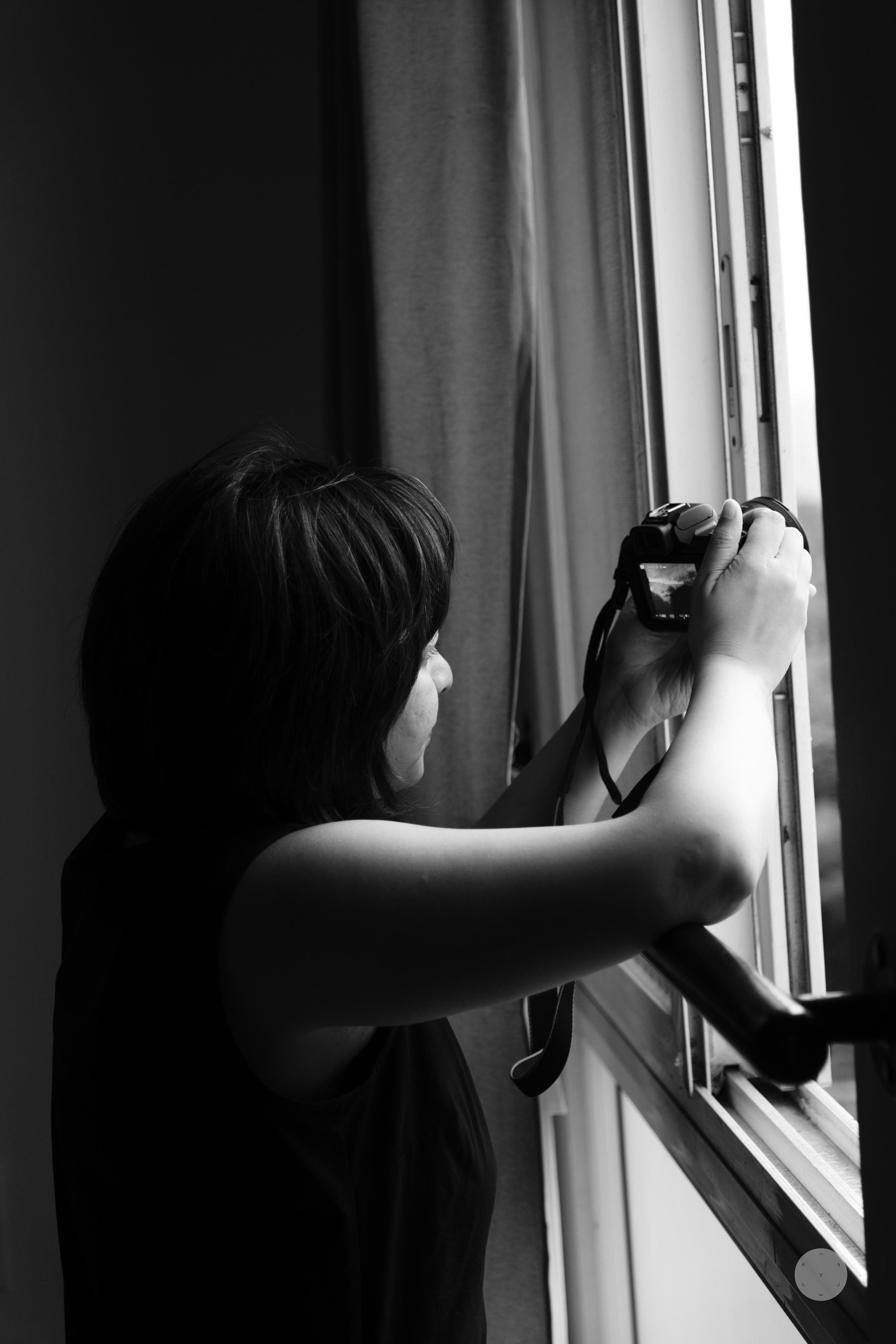 A person with short dark hair is taking a photo with a camera near a large window, with the sky and buildings visible outside.