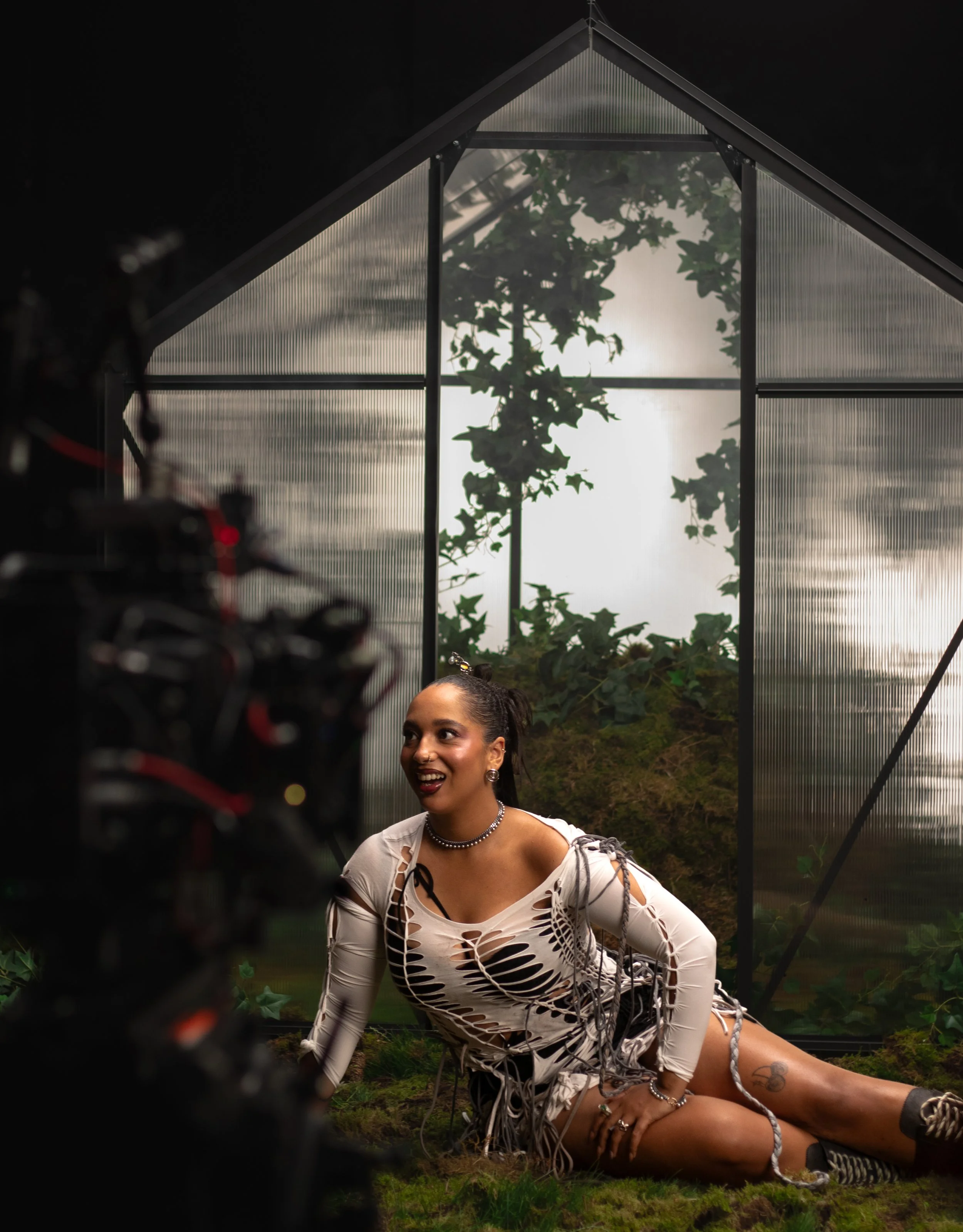 A woman with dark hair and a nose ring is smiling looking at the camera in between shots. She is outside a greenhouse transparent roof and walls, with plants and greenery. She is wearing a ripped, long-sleeve dress with a hole pattern.