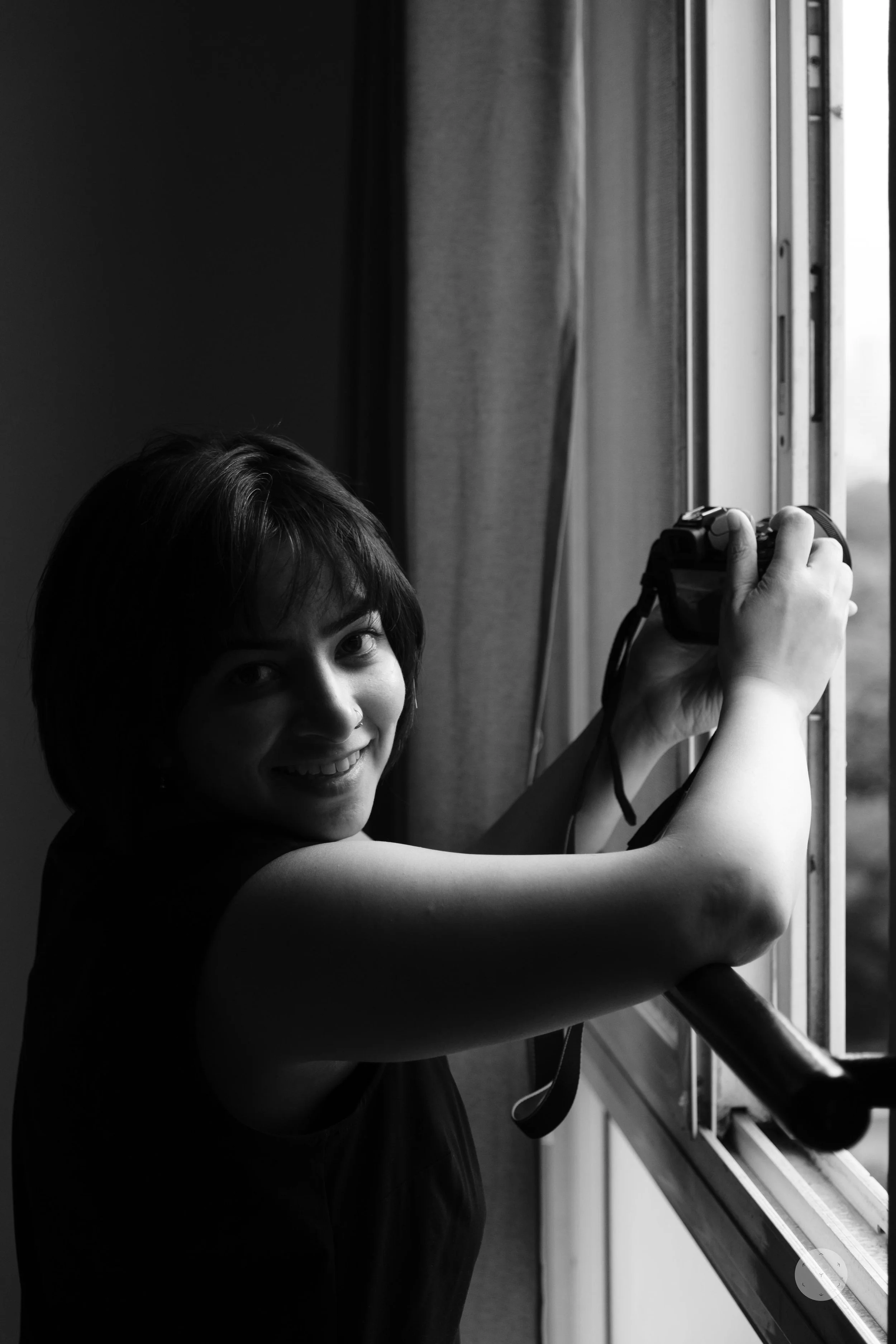 A woman with short dark hair smiling while holding a camera near a window.