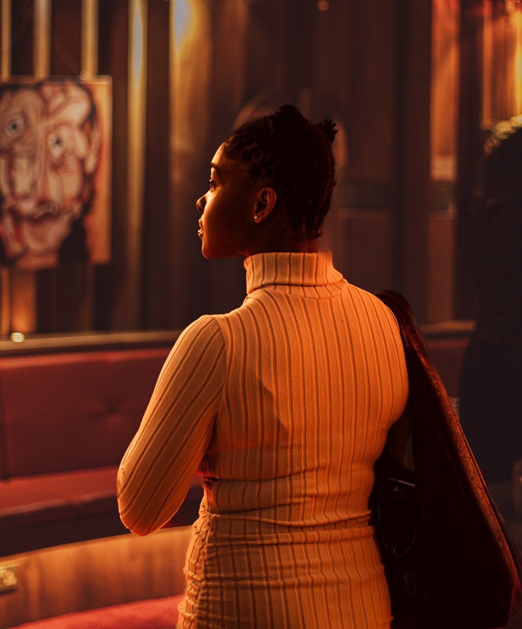 A woman with short, natural hair wearing a beige ribbed turtleneck top, standing and looking at art in a gallery in a dimly lit room.