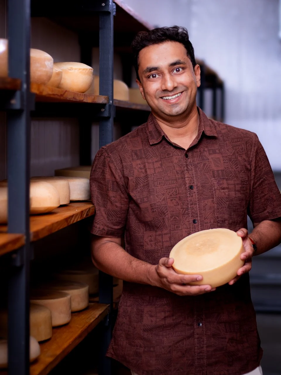 A man smiling and holding a large wheel of cheese in a cheese storage room with shelves filled with cheese wheels.