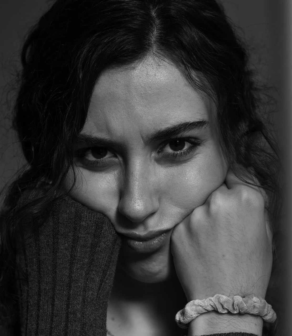 A close-up black and white portrait of a woman with wavy dark hair, looking directly at the camera with a slight smile. Her face is resting on her hand, which has a scrunchie around the wrist.